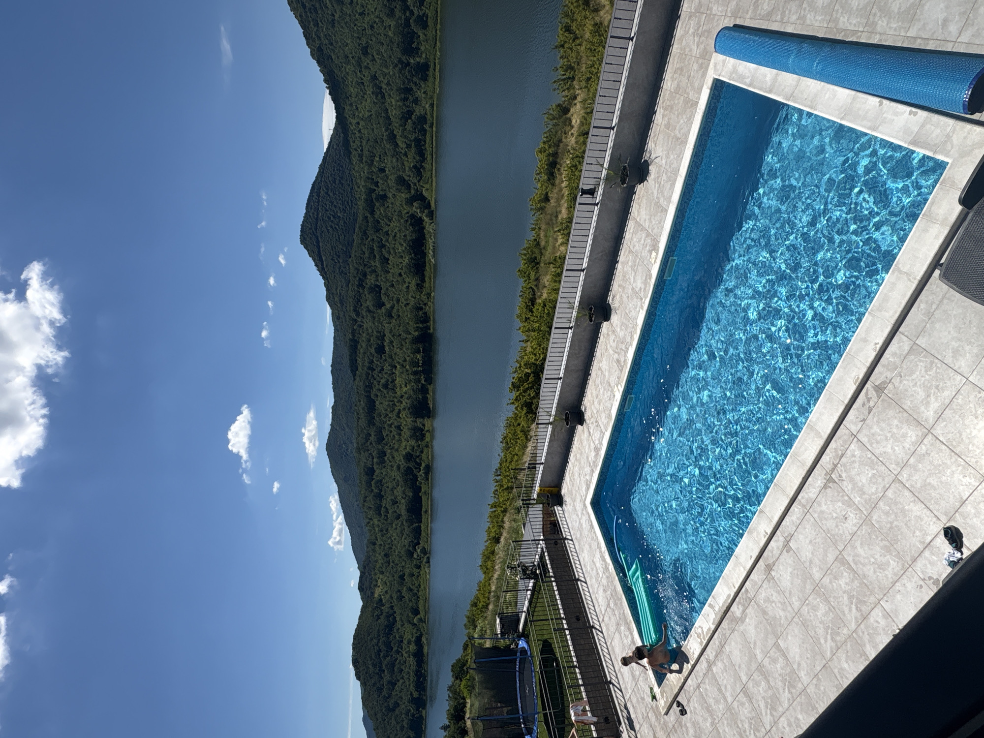 The image shows an outdoor swimming pool with bright blue water, tiled surround, and a pool cover. Two young boys are in or near the pool; one is standing at the edge with a green inflatable raft nearby, and the other is in the water. A trampoline is visible in the background to the left, with a metal fence bordering the property. Beyond the property, there is a large body of water, likely a lake, with lush green forested hills and mountains rising behind it under a bright blue sky with scattered white clouds. The overall scene suggests a recreational area in a natural, scenic environment.
