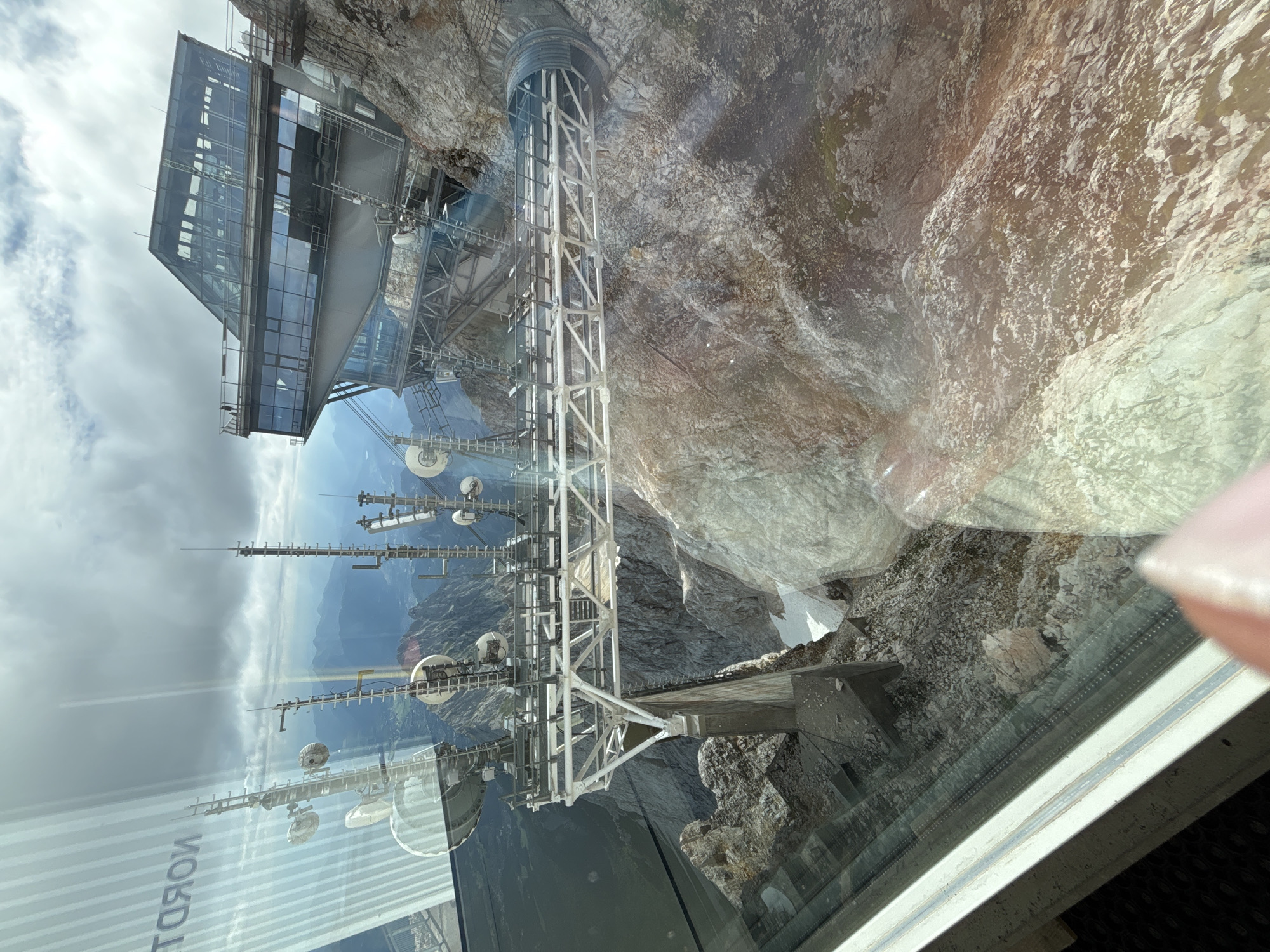 The image shows a view from a window looking out at a mountainous landscape. In the foreground, a modern glass building with angular lines is perched on the side of a rocky mountain. A metal structure, possibly a walkway or part of a communication tower, extends from the building over a steep drop. Various antennas and satellite dishes are mounted on this structure. The background reveals a panoramic view of rugged, grey mountains under a cloudy sky. The reflection on the glass in the foreground shows vertical stripes and what appears to be text, possibly from an advertisement or sign. The overall scene suggests a location at a high altitude, likely a tourist attraction or a research facility in a mountainous region. The image is taken from inside a moving vehicle, likely a cable car, given the slight blur and the perspective. The mention of Ehrwald, Austria, suggests this location could be a prominent peak or viewpoint accessible from the town.