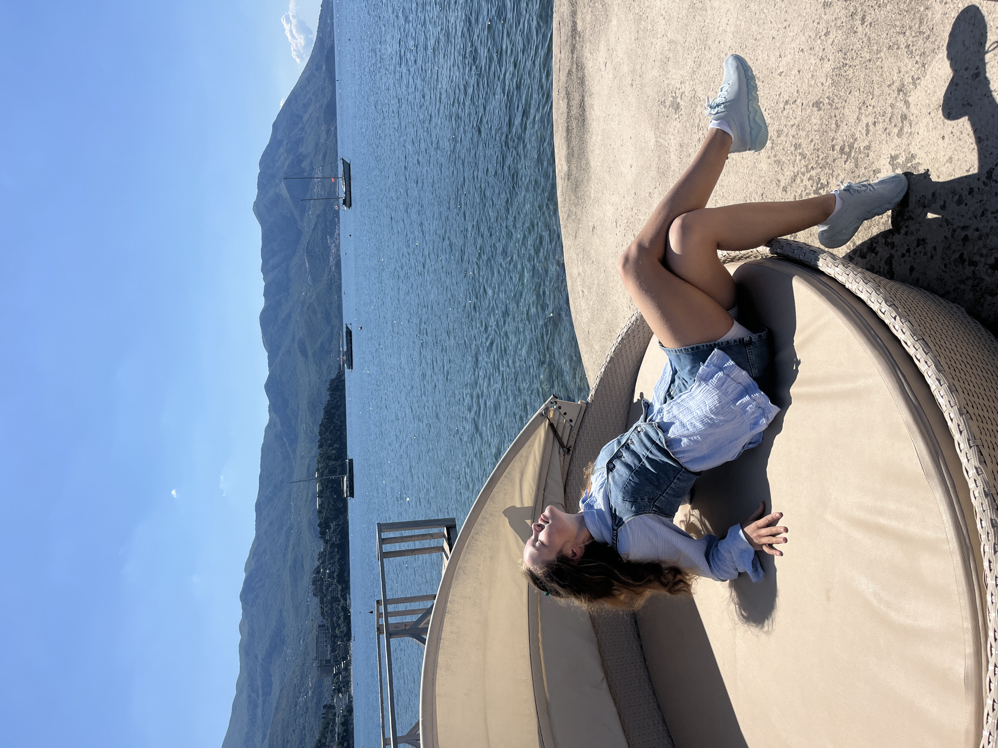 A young woman wearing light-blue shoes, denim shorts, a blue blouse, and a denim overall top, lies on a beige round wicker sunbed, resting on a concrete surface next to water. The woman is facing the camera, with her legs angled upwards and bent at the knees. Behind her, a body of water stretches towards a mountainous terrain, with several boats visible on the water. The sky is a clear, light blue. The scene appears to be taken in a coastal location, similar to Budva, Montenegro.