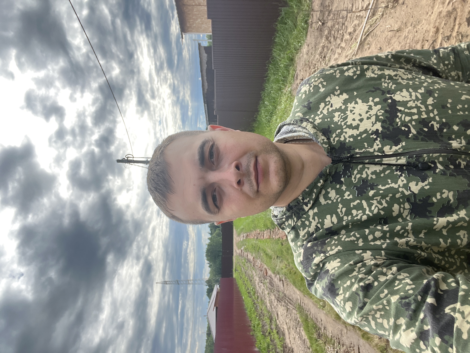 A man in a camouflage hooded jacket stands outdoors in what appears to be a rural area, possibly the village of Razdolye, Russia, based on provided context. The man is facing forward, with the camera positioned at a slight upward angle. He has short brown hair and fair skin, and is looking directly at the camera. Behind him, a dirt road or path runs lengthwise, flanked by green grass and dark brown metal fencing on either side. In the background, there are houses, trees, and a tall transmission tower visible against a cloudy sky. The ground is uneven and appears to be dirt and some gravel.