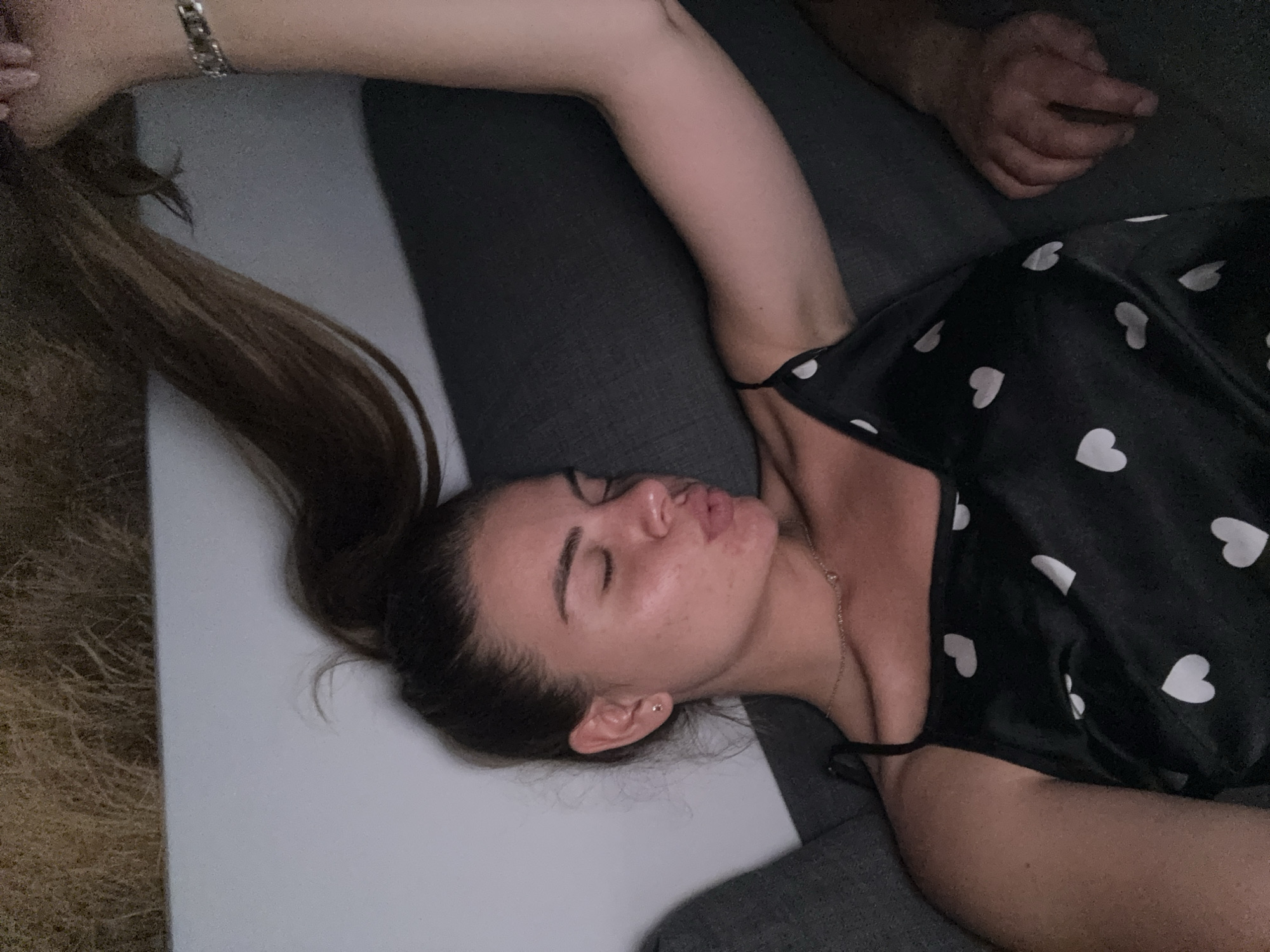 The image shows a woman lying on her side on a dark gray couch. She is wearing a black slip with a pattern of white hearts. Her eyes are closed, and her lips are pursed as if she is about to kiss someone or something. Her right arm is raised above her head, and her hair, which is pulled back in a ponytail, is draped over the back of the couch. Her left arm is bent at the elbow, resting on the couch. A person's hand is visible on the couch to the right of the woman. The background is blurred, but it appears to be an indoor setting with some light-colored furniture or wall and a textured, possibly grassy, surface above. The image does not provide any information that would allow for its localization in the city of Plovdiv, Bulgaria.
