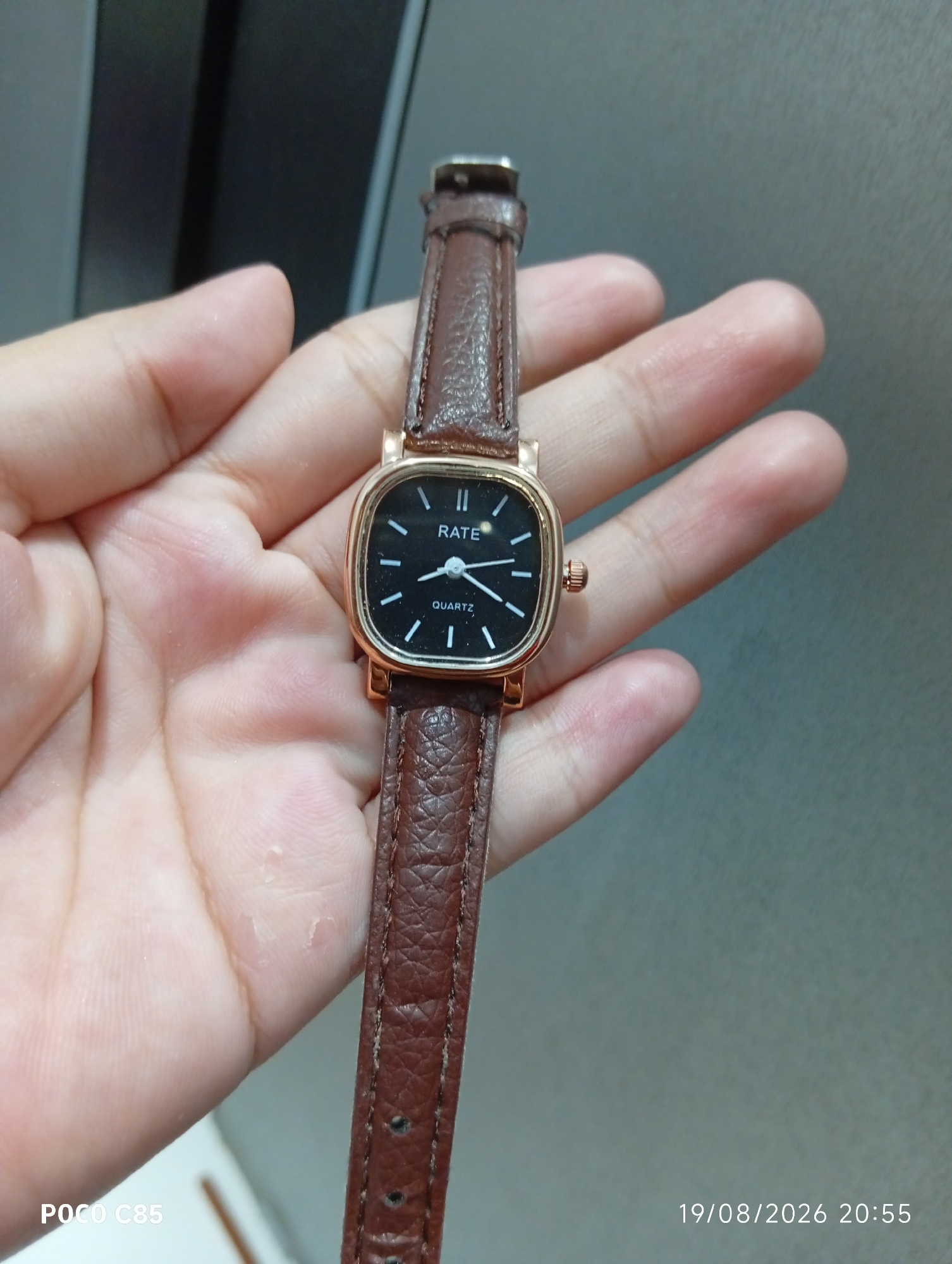 A close-up, eye-level shot shows a person's right hand holding a square-faced, gold-toned watch with a brown leather band. The watch face is black with white hour markers and hands, and the brand name "RATE" is visible at the top, with "QUARTZ" below it. The watch band is textured and appears worn. The background is a blurred, neutral gray wall. A timestamp in the bottom right corner reads "19/08/2026 20:55". A watermark "POCO C85" is present in the bottom left corner. The setting is likely indoors in South Tangerang, Indonesia, given the watermark.