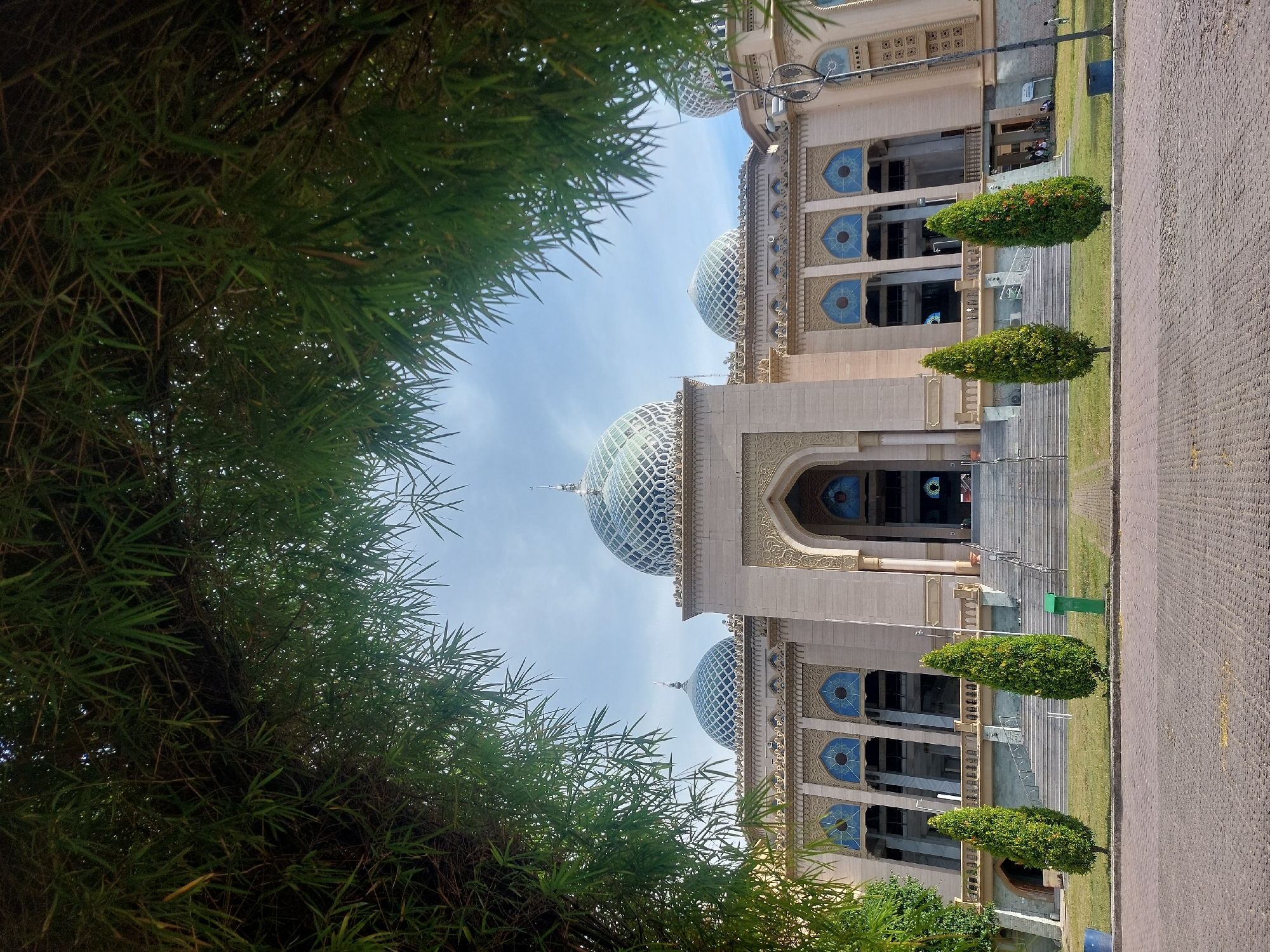 A wide-angle vertical shot shows the front of a mosque, framed by the branches and leaves of a bamboo tree. The mosque features a grand entrance with a wide staircase leading up to a large arched doorway. The building is constructed with light-colored stone and adorned with intricate geometric patterns and blue stained-glass windows. Several domes, covered in a diamond-shaped pattern, crown the mosque. In front of the mosque, a paved area leads to a grassy lawn where several manicured evergreen trees are planted. The sky above is cloudy and pale blue. There are no people clearly visible in the immediate foreground, but a few figures can be seen on the grass near the mosque's right side. This is the Baiturrahman Grand Mosque in Lhokseumawe, Indonesia.
