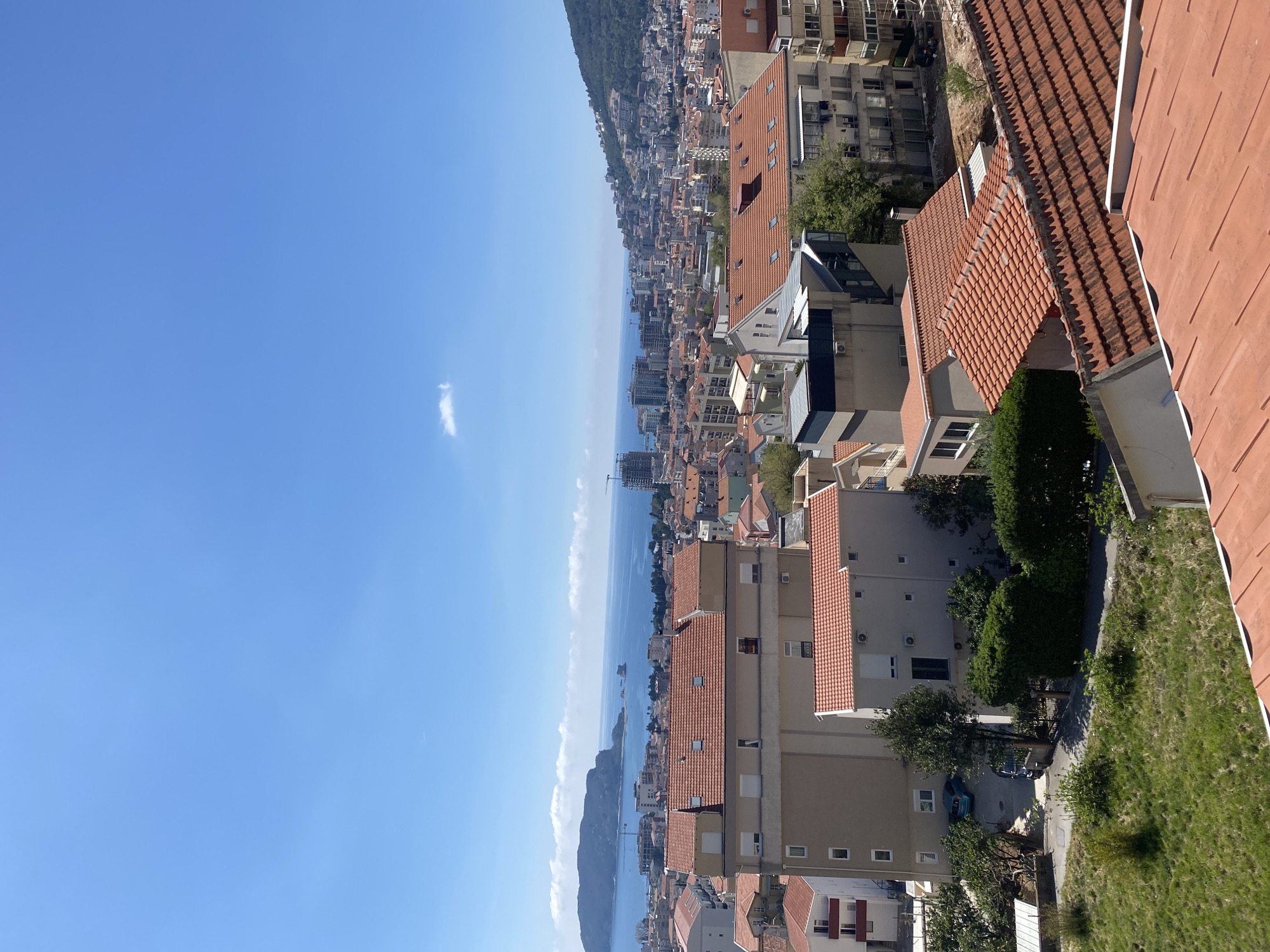 The image is an elevated view of Budva, Montenegro, showing a cityscape with reddish-tiled roofs of various buildings extending down to the Adriatic Sea. In the distance is a mountainous coastline. The sky is blue with minimal clouds. Some buildings are close to the viewer and appear as residential structures, while the buildings become more dense as they descend towards the sea. A patch of grass is visible on the lower right.