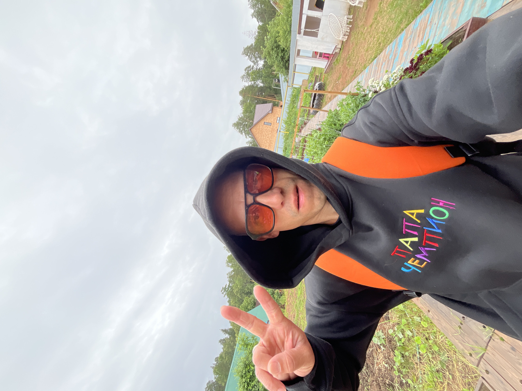This is a selfie of a person wearing a black hoodie with an orange backpack strap visible. The hoodie has colorful embroidered text on the chest that reads "ПАТТА ЧЕМПИОН". The person is wearing sunglasses and is making a peace sign with their right hand.
The background appears to be a rural or suburban outdoor setting. There are several buildings visible, including a house with a brown roof and a partially constructed building with white walls. A wooden pathway or deck is present, bordered by greenery and some low-lying plants with dark red and white flowers. Tall trees and lush green foliage surround the area, suggesting a natural environment.
The sky is overcast and cloudy, indicating a possibly cool or damp day. The lighting is diffused, suggesting it's daytime, but not necessarily sunny. The overall impression is of a casual, possibly vacation or leisure setting in Chaikovsky, Russia. The person's expression and peace sign gesture convey a sense of lightheartedness or positivity.