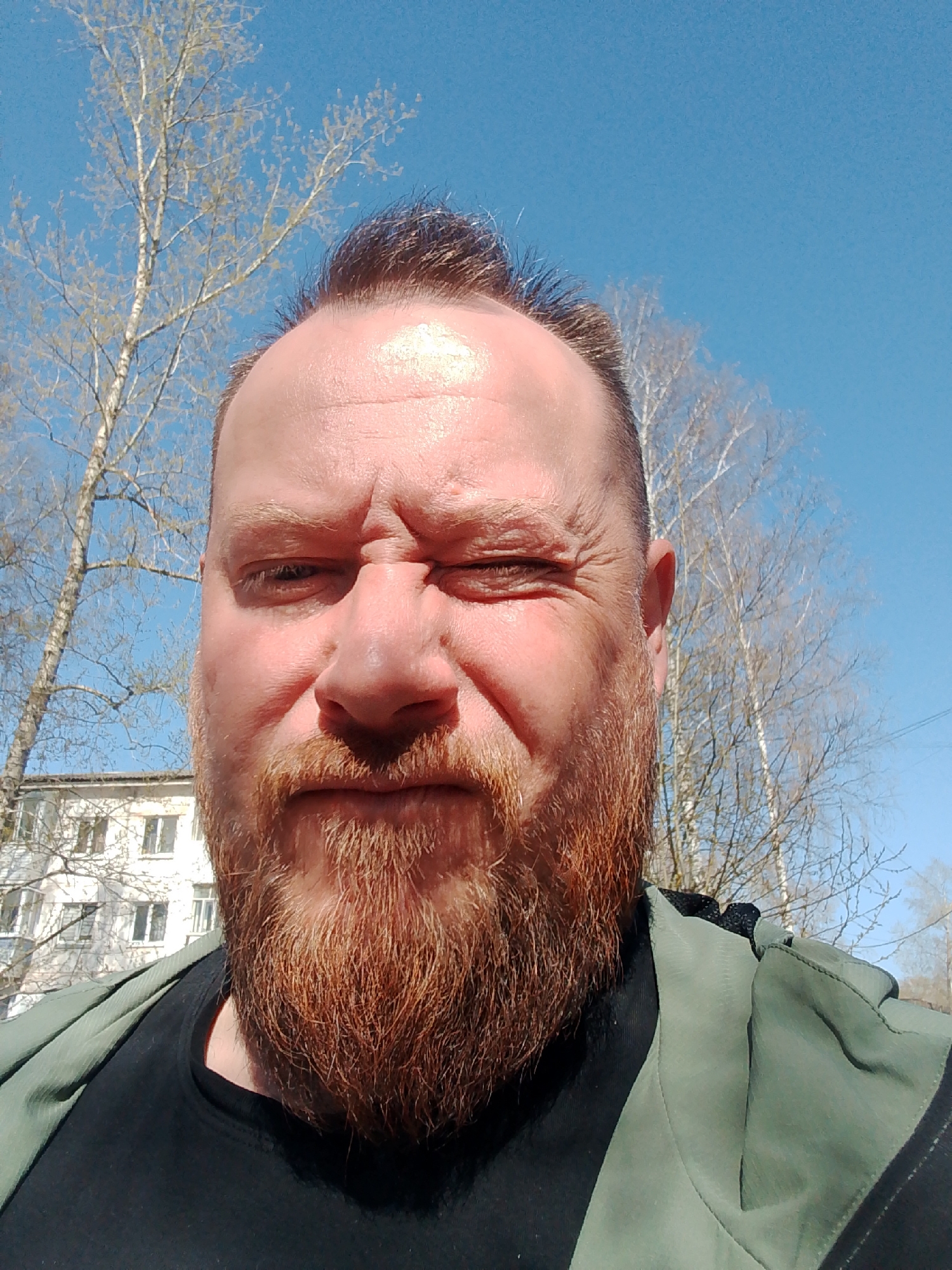 A selfie depicts a middle-aged Caucasian man with a red beard, wearing a black t-shirt and an olive-green jacket. He is squinting and wrinkling his forehead. Behind him, deciduous trees with sparse branches are visible against a bright blue sky. In the lower-left corner, a multi-story building with windows is partially visible. This scene takes place in the city of Chaikovsky, Russia.