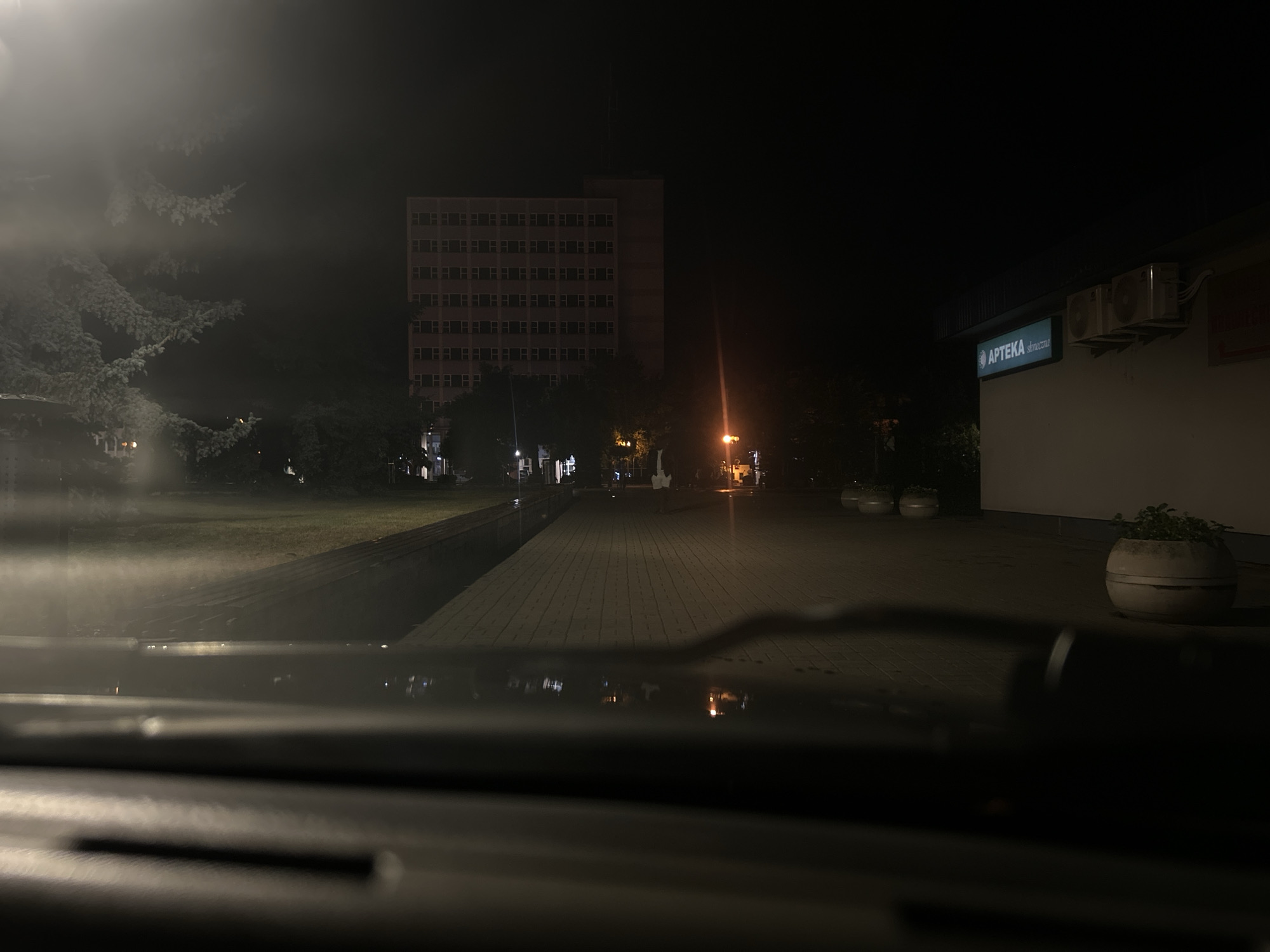 The image depicts a nighttime scene, likely taken from inside a vehicle, looking out onto a street or public area in Siedlce, Poland. In the background, a tall, multi-story building with numerous windows is visible, appearing to be a residential or office building. To the right, a building with a sign that reads "APTEKA" (Polish for "pharmacy") is illuminated, with an outdoor air conditioning unit mounted on its wall. The foreground shows a dashboard or hood of a car, with reflections of lights and the environment. The ground in front of the buildings is paved with interlocking bricks, and there are several round planters, some containing greenery, along the walkway. Trees are present, some of which are illuminated by artificial lights, creating a slightly hazy or misty atmosphere in certain areas, possibly due to fog or moisture. The overall impression is a quiet, urban setting at night.