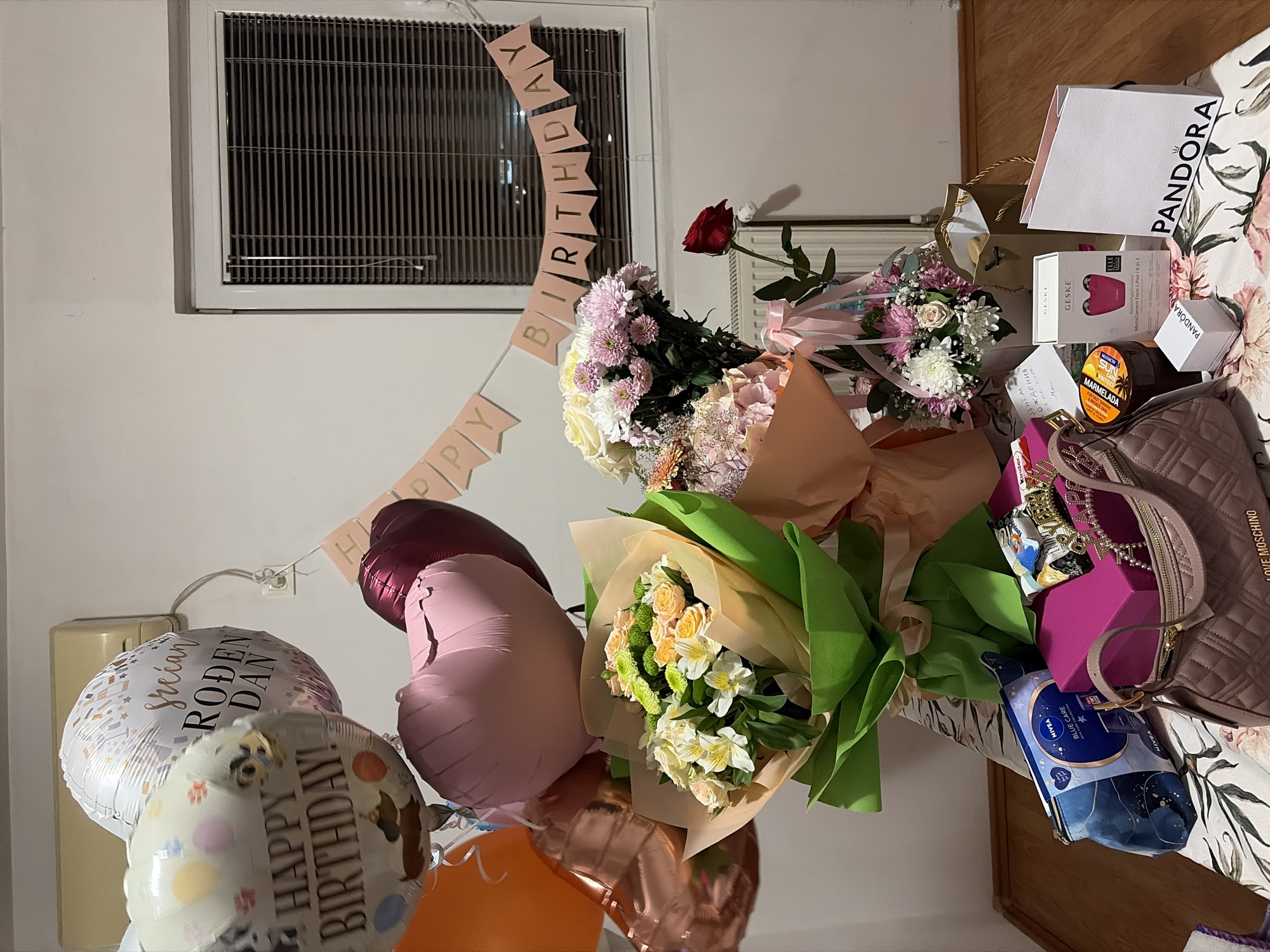 The image depicts a domestic interior setting, likely a bedroom or living room, with a wooden floor and white walls. A window with closed horizontal blinds is visible on the right side. The scene is filled with birthday decorations and gifts.
Dominating the upper portion of the image are several balloons: a white one with confetti and text in a Slavic language that appears to say "Srećan rođendan" (Happy Birthday), a cream-colored balloon with various dog breeds and the text "HAPPY BIRTHDAY!", an orange balloon, and two heart-shaped balloons, one pink and one dark red. A pink banner spelling out "HAPPY BIRTHDAY" is strung across the wall.
In the foreground, several bouquets of flowers are arranged. The largest bouquet features a mix of white, yellow, and green flowers, wrapped in green and tan paper. Another bouquet to its right contains pink and white chrysanthemums and other delicate flowers, with pink ribbons. A single red rose is placed in a vase to the right of the flower arrangements.
On a patterned tablecloth or bedspread, a collection of gifts is displayed. These include a pink quilted handbag with a "LOVE MOSCHINO" logo, a blue and black cosmetic bag with "NIVEA BLUE CARE" branding, a bright pink gift box, and several smaller items such as a jar of what appears to be honey, chocolates, a small white box with "PANDORA" branding, a larger white "PANDORA" shopping bag, and a box for a "GESKE SmartMicroCurrent Face-Lifter". A small, ornate crown is also visible among the gifts.
The overall impression is a festive and celebratory arrangement of birthday gifts and decorations, presented in a domestic environment. There are no people visible in the image. The context of Budva, Montenegro, is not deducible from the visual elements within the image itself.