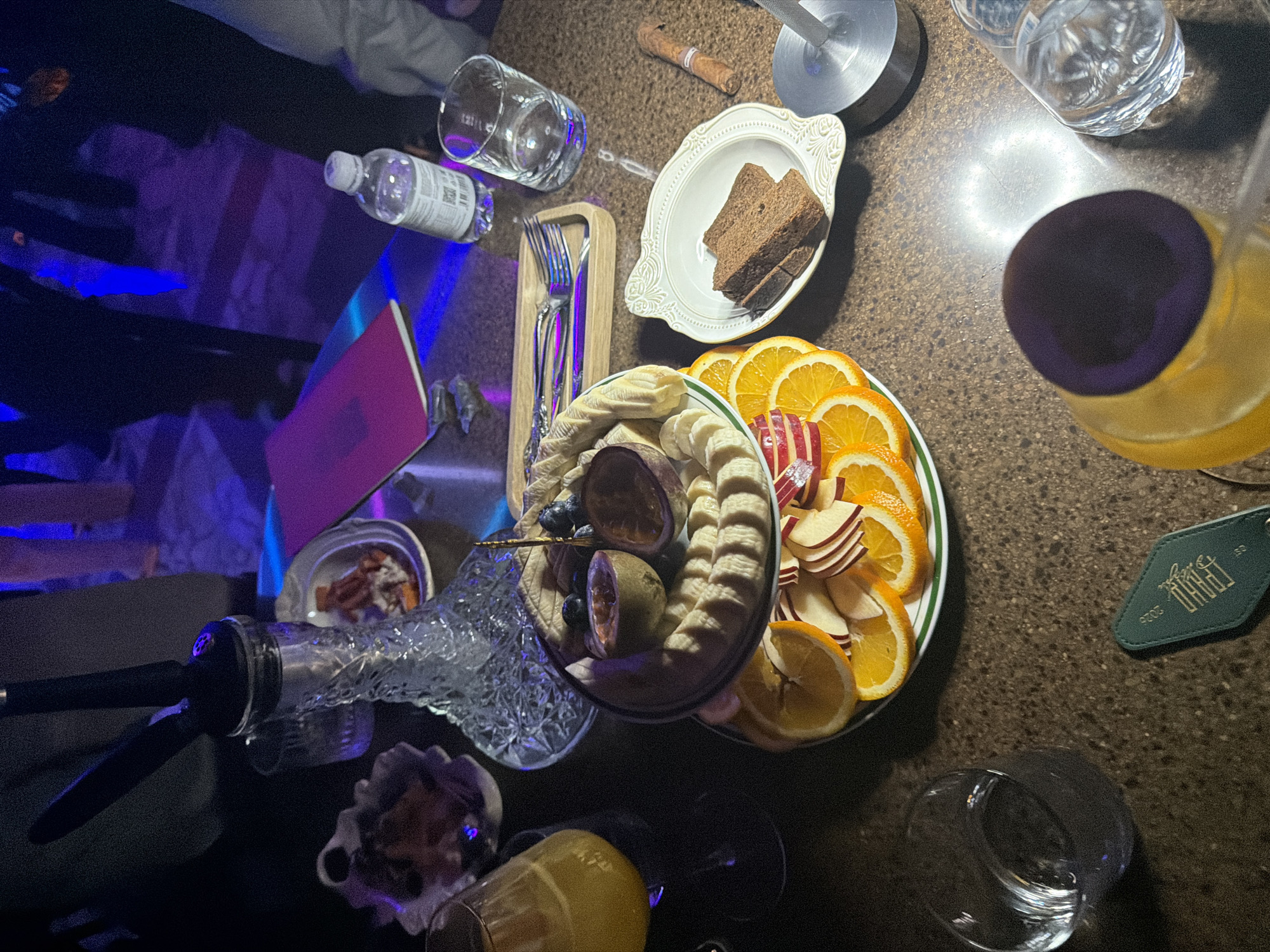An overhead view captures a table laden with food and drinks in what appears to be an evening social gathering in Krasnoyarsk, Russia. The scene is dimly lit by artificial blue and purple lights, creating a relaxed, lounge-like atmosphere.
The table, with its dark, speckled surface, holds a variety of refreshments. Food items include a large white plate with neatly arranged slices of oranges and red apples, and another plate featuring sliced bananas, blueberries, and halved passion fruit. A smaller white dish contains two slices of dark bread, while a small bowl holds unidentifiable brown morsels, possibly nuts or dried fruit.
Beverages consist of a plastic water bottle with a visible QR code and unreadable text, several clear glasses (some empty, some holding water), and a large pitcher filled with an orange or yellow liquid, likely juice, topped with a dark lid. Another glass also contains a yellowish drink.
Other notable objects contributing to the social context include a glass hookah with a black hose, partially visible on the left, suggesting a leisure activity. A wooden tray holds two forks. A small, dark pink or purple book or journal is open on the table. A rolled-up cigar or tobacco stick is also present. A distinctive dark green keyring or tag is visible, embossed with the white text "ПИВУ 2020" and the number "50", possibly indicating a brand, year, or item. The overall impression is a casual, comfortable gathering.