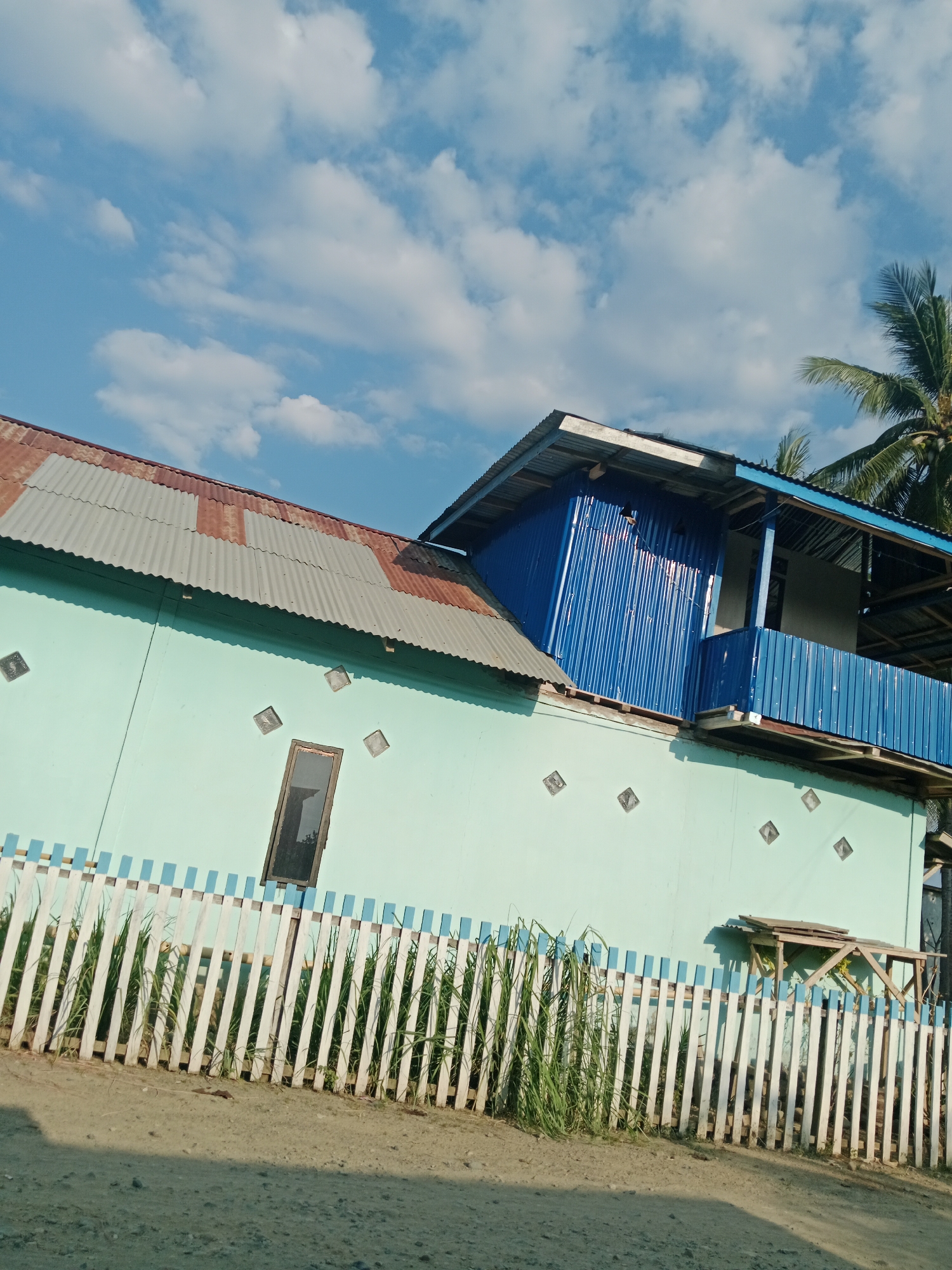 The image depicts a side view of a two-story building with a light blue facade, located in Lakea Satu, Indonesia. The lower story has a corrugated metal roof with patches of rust and a light blue and white striped wooden fence in front of it. Tall green grass grows behind the fence. There is a narrow, dark rectangular window on the lower story, surrounded by four diamond-shaped decorative elements. The upper story is painted in a vibrant blue, featuring a balcony with a blue metal railing. A palm tree is visible on the far right, suggesting a tropical environment. The sky is a clear blue with scattered white clouds. The ground in front of the fence is a dirt road.