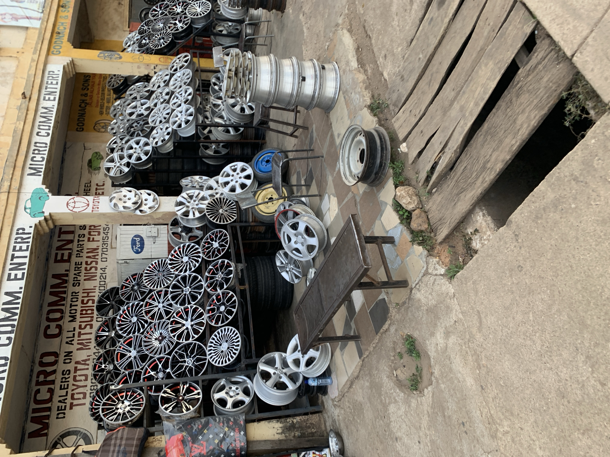 The image shows the exterior of a business called "MICRO COMM. ENTERP." located in New Stadium, Nigeria, as indicated by the phone number area code. The business is a dealer in motor spare parts, specifically catering to brands like Toyota, Mitsubishi, Nissan, and Ford. The shop is filled with various car rims and wheels, stacked on racks and displayed in front of the building. Next door is GODNACH & SONS, dealers in new wheels, batteries and wheel balancing. A wooden-covered drainage or opening is visible on the side of the shop. The street is paved with concrete. There are no people visible in the image.