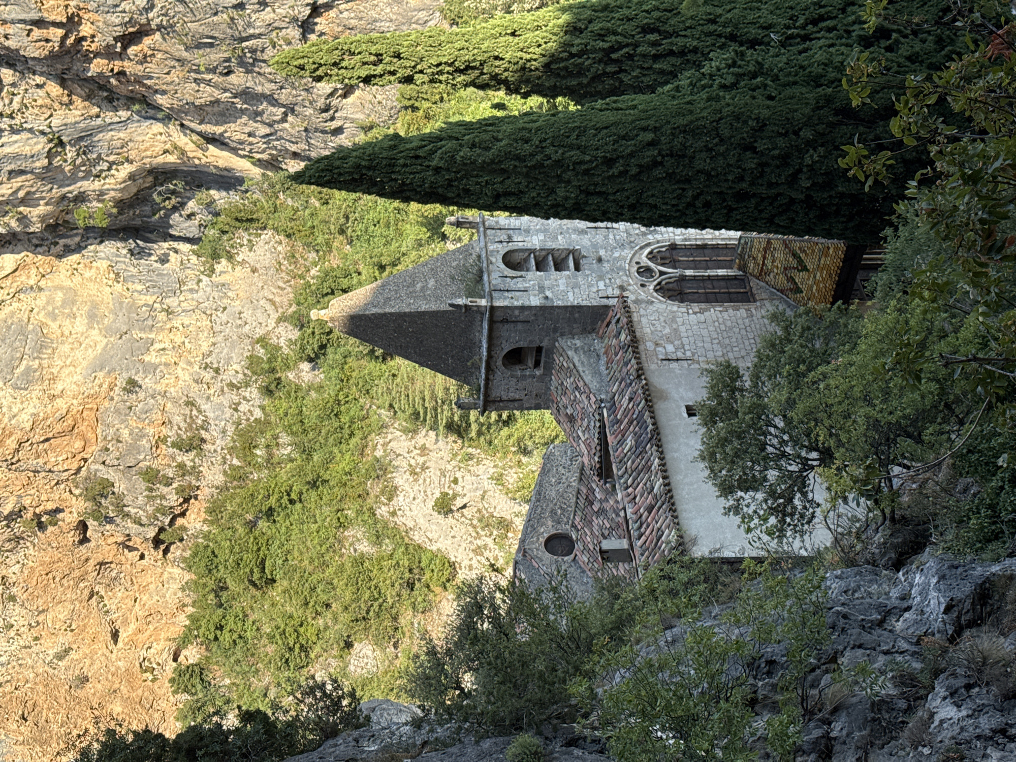 This image depicts a stone church with a tiled roof and a steeple, nestled within a rugged, rocky landscape covered in green foliage. The church is partially obscured by trees, with tall, slender cypress trees flanking the right side of the steeple. The background consists of a steep, rocky cliff face with patches of vegetation. The scene appears to be illuminated by natural light, casting shadows and highlighting the textures of the stone and rock. The architectural style of the church, with its arched windows and stone construction, suggests it is an old building. The overall impression is one of a remote, historical structure integrated into a natural, mountainous environment. The location is identified as Moustiers-Sainte-Marie, France, a village known for its dramatic setting within a gorge.
