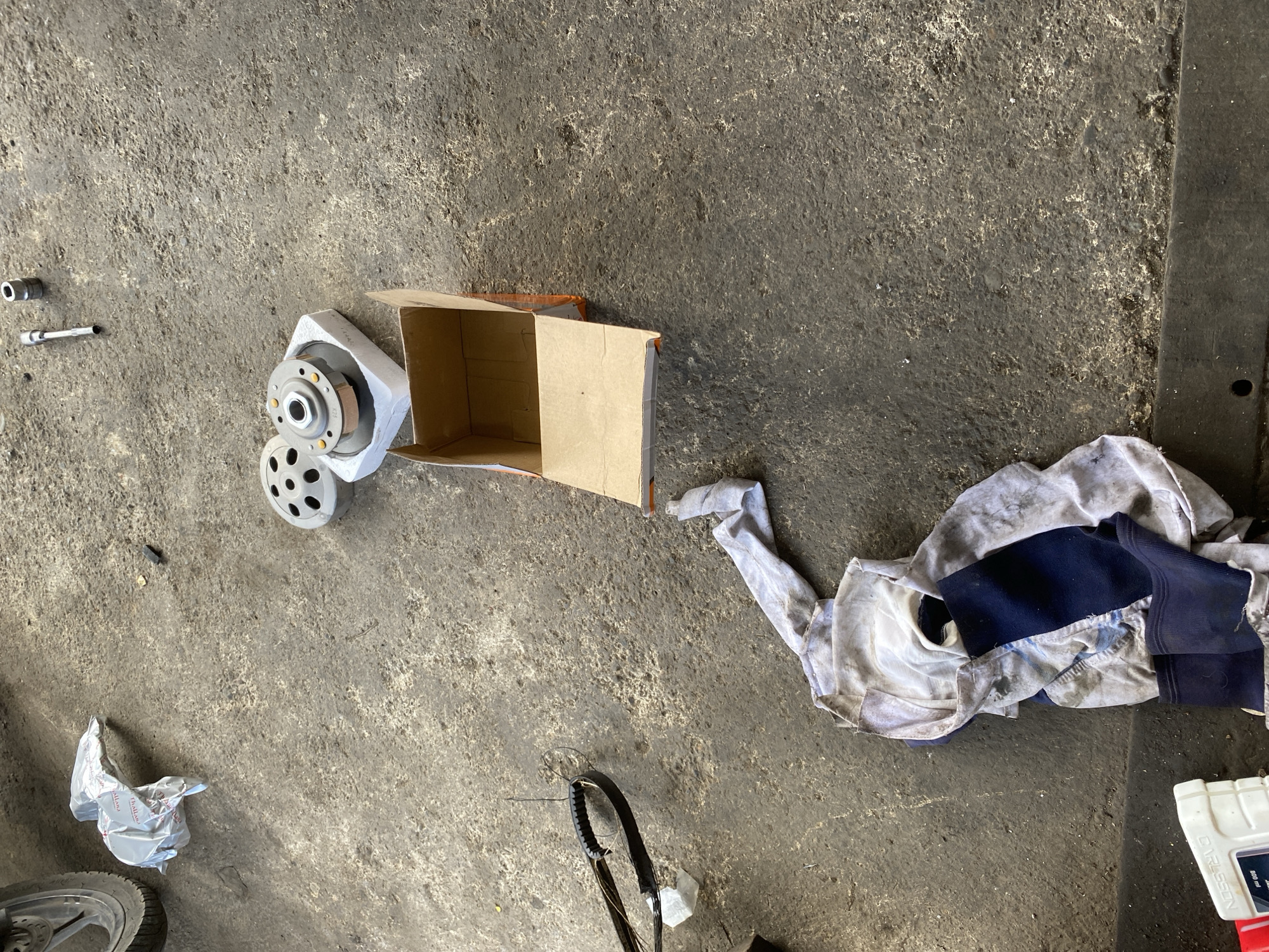 An overhead, eye-level, medium shot shows a concrete garage floor with various motorcycle parts and tools scattered about, suggesting a repair or maintenance area. A motorcycle wheel is visible on the left edge of the frame. Several metal parts, possibly clutch components, are arranged near a partially opened cardboard box in the center of the image. A black rubber belt, likely from a motorcycle, lies to the left of the box. A soiled cloth or rag is draped on the floor in the lower center of the image, and a white plastic bottle, possibly containing lubricant or oil, is positioned in the lower left corner. The scene appears to be set in an urban environment, implied by the concrete floor and the nature of the objects present.