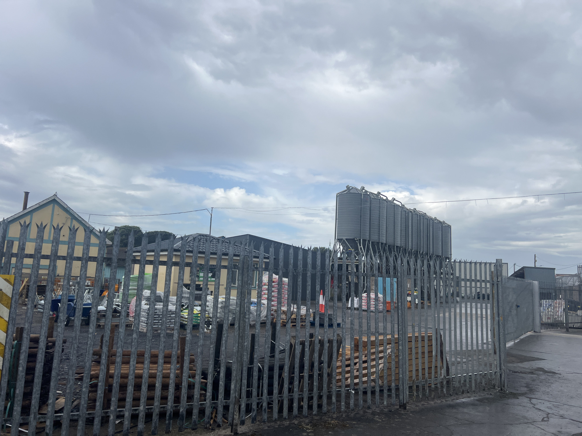 The image shows an outdoor storage area with a grey metal fence in the foreground. Behind the fence, there are stacks of bagged materials, likely for construction or agriculture, on pallets. A blue wheelbarrow is visible near some of the stacks. In the background, there are large industrial silos and buildings. The sky is cloudy and overcast. The ground is paved with asphalt.