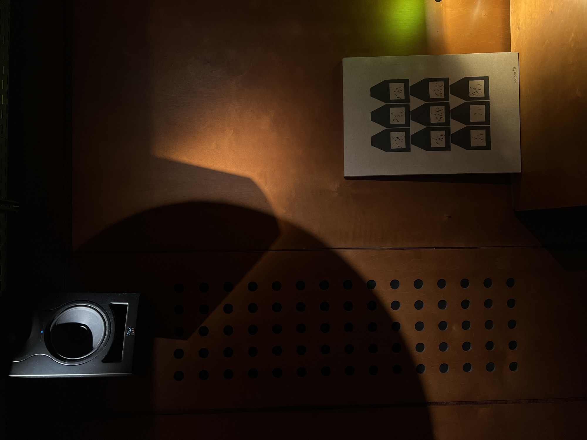 This media file presents a dimly lit interior scene, likely an indoor space in Yerevan, Armenia, characterized by warm, reddish-brown wooden walls. The setting has a modern and somewhat artistic aesthetic.
The primary subjects include a piece of wall art, an electronic device, and a perforated wall panel. On the upper right portion of the wooden wall, a light-colored, rectangular artwork or poster is displayed. This piece features a stark black and white design: a grid of nine identical, stylized shapes resembling truncated pyramids or beakers, each containing an irregular, speckled pattern. Along the right edge of this artwork, the vertical text "nununu '21" is visible.
In the lower-left corner, a sleek, black electronic device, which appears to be a speaker or a small subwoofer, is visible. It features a central circular driver, a small glowing blue LED indicator light, and a stylized logo resembling "PS" or "PA" on its right side.
Below the artwork and extending towards the lower right, a section of the wooden wall is covered by a panel with a uniform grid of small, circular perforations, possibly serving as an acoustic element or decorative screen.
The scene is defined by its dramatic lighting. A strong, unseen light source casts sharp, angular shadows across the wooden surfaces, creating distinct geometric patterns and enhancing the depth of the space. There's a subtle yellowish-green light originating from the upper left, adding a cool contrast to the warm tones of the wood. The overall ambiance is static and moody, with no human activity taking place. The time of day is not explicitly clear but the controlled, artificial lighting suggests an indoor setting, possibly at night.