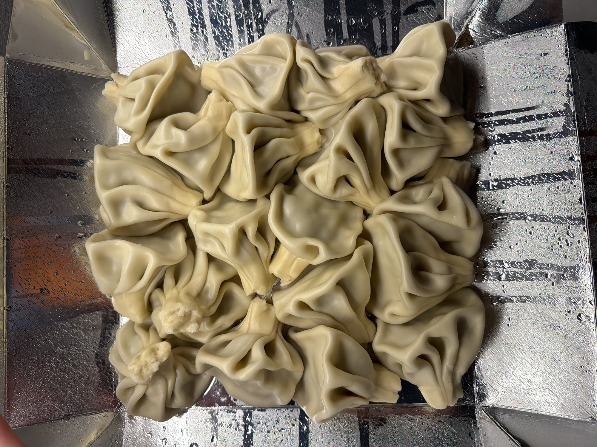 A close-up shot shows a collection of boiled dumplings, likely khinkali, arranged on a silver, metallic surface within a square container. The dumplings are light beige with numerous folds and twisted tops. Some dumplings appear to have a small portion of the filling exposed. The silver surface is also wet with water droplets. The photo may have been taken in Tbilisi, Georgia, as khinkali is a common Georgian dish.