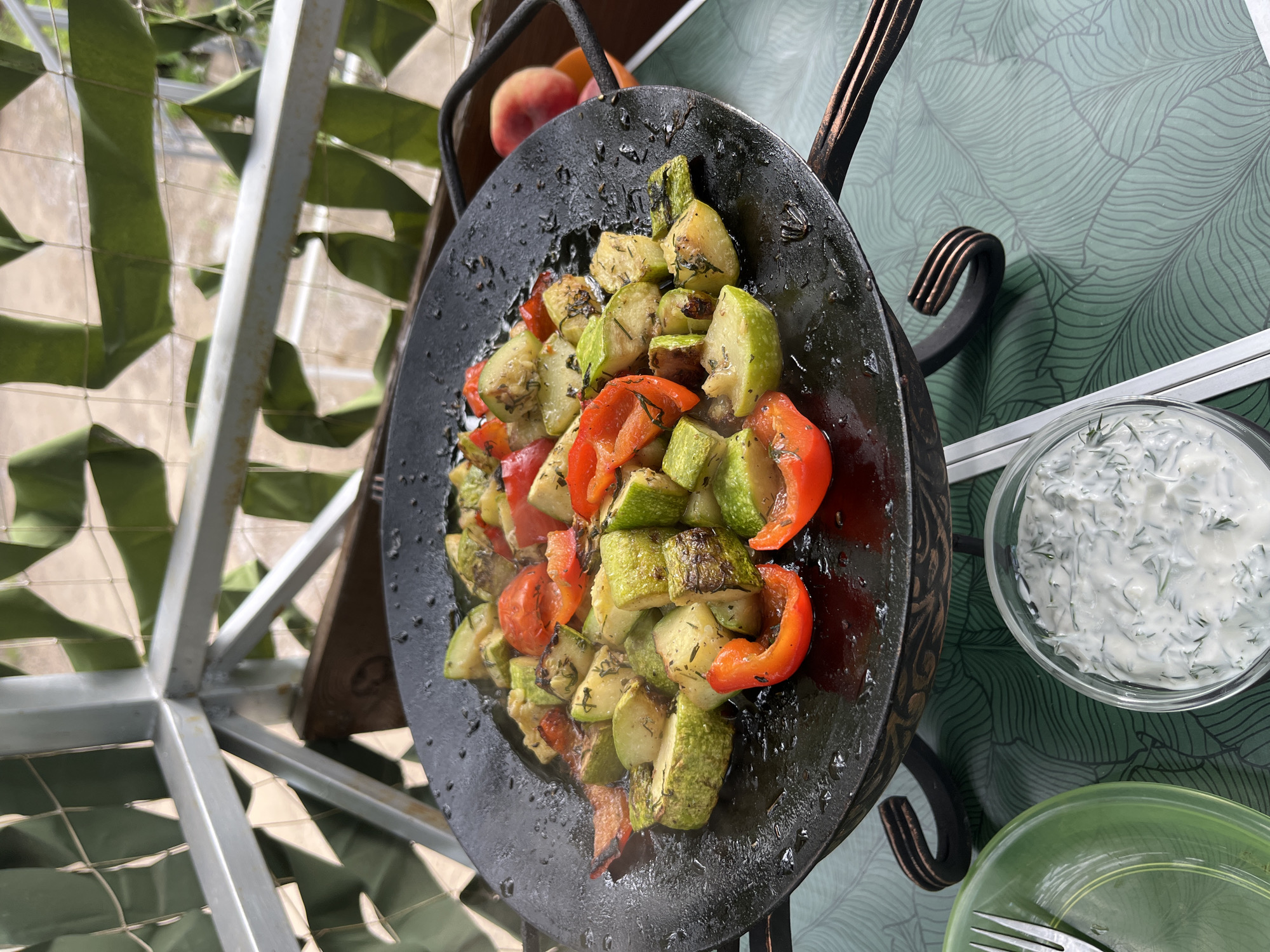 An overhead shot shows a black, shallow cooking pan filled with sautéed zucchini, red bell peppers, and herbs. The pan is resting on a decorative metal stand. In the foreground, a glass bowl holds a white dip with green herbs, and a green plate with a fork sits to the left. The table has a green, leaf-patterned surface. In the background, a metal structure with green fabric draped over it forms a decorative screen, and a peach is visible in an orange bowl. The scene is outdoors on a balcony or patio. This image does not provide enough visual information to determine its location in Chaikovsky, Russia.