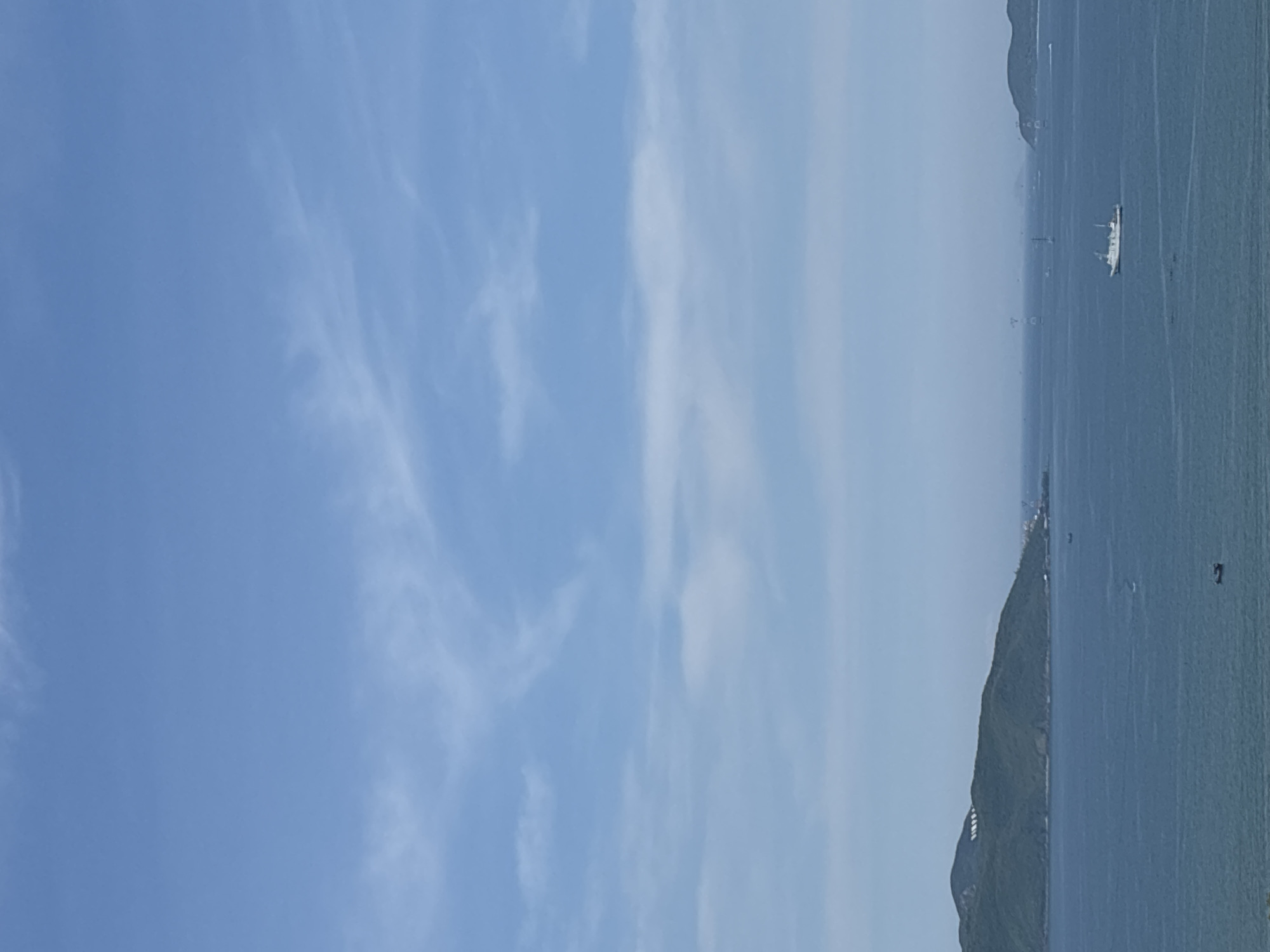 A high-angle, wide shot captures a serene coastal landscape under a clear blue sky with wispy clouds. In the foreground, a vast expanse of deep blue water stretches towards the horizon. A single white boat navigates the tranquil sea, positioned slightly to the right of the frame's center. In the midground, a lush, green, hilly island dominates the left side of the image, its slopes dotted with white structures. Further in the distance, other smaller islands and landmasses are faintly visible, blurring into the hazy horizon. The overall scene evokes a sense of calm and open space.