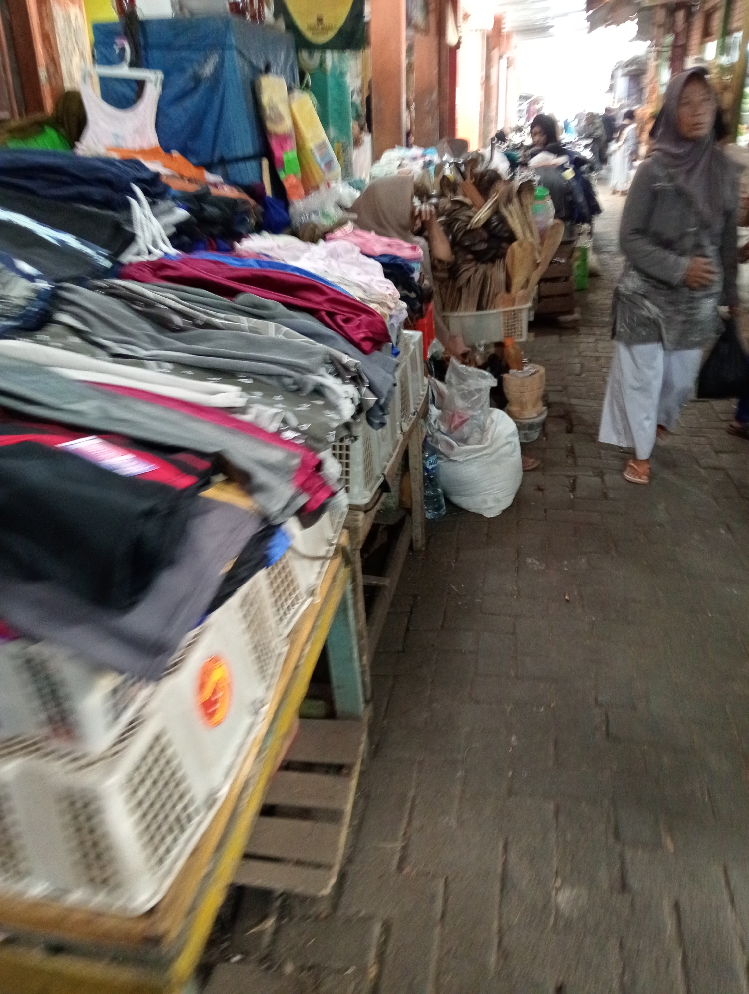 The image depicts a street scene in Grabag, Indonesia, likely a market or shopping area. In the foreground, a stall displays a variety of clothing items, including t-shirts, pants, and possibly blankets, neatly folded or hung on display racks. The textiles are a mix of colors and patterns, with a focus on casual wear. The stall itself is constructed with wooden elements and utilizes white plastic crates for organization.
Further back, several individuals are visible. One woman, wearing a grey hijab and a grey top over white pants, walks away from the camera. Another woman, also in a hijab, is seen partially obscured by a display of wooden utensils and what appear to be dried goods. Other people are present in the background, some walking, others possibly browsing. The environment suggests an outdoor or semi-covered market with paved flooring. The overall impression is one of a bustling marketplace with goods for sale and people engaged in shopping activities.