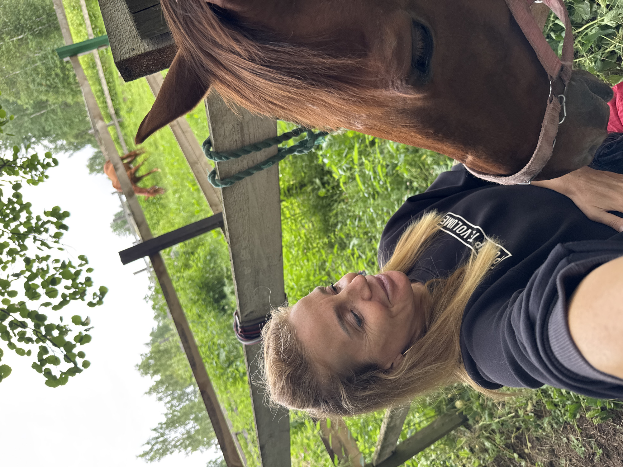 The image shows a woman and a horse outdoors in a grassy area. The woman is wearing a black hoodie and has blonde hair. She is looking at the horse, which has a brown coat and is wearing a bridle. The horse is leaning in towards the woman, and their faces are close together. The scene appears to be taken during the daytime, with natural light illuminating the subjects. The background consists of green grass, trees, and a wooden fence. There is some text visible on the woman's hoodie, but it is partially obscured and difficult to read. The overall mood of the image suggests a moment of connection and affection between the woman and the horse. The location context provided is Mars, Russia.