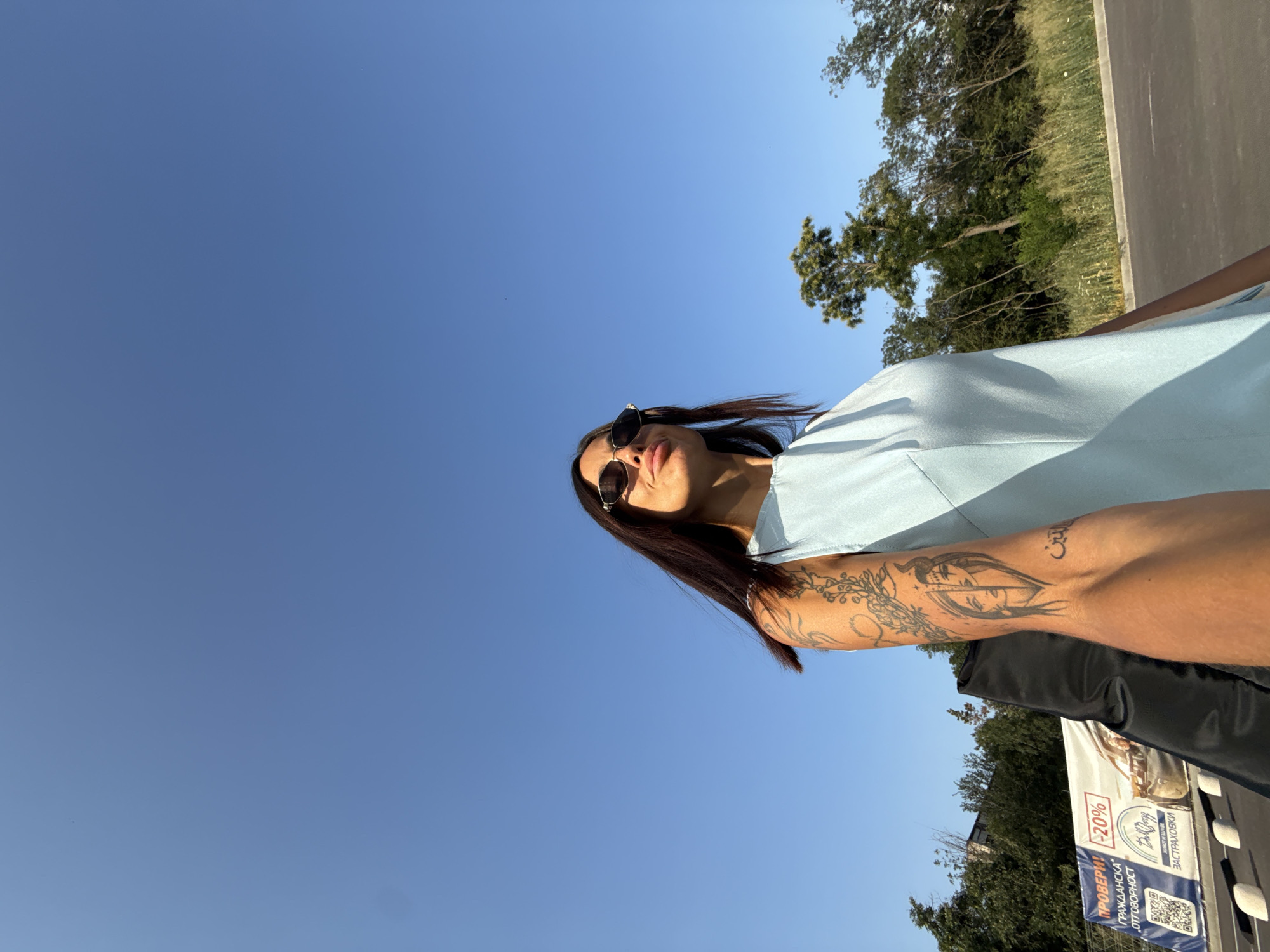 A woman with dark hair, sunglasses, and a light blue dress stands outdoors under a clear blue sky. She has tattoos on her left arm. Behind her, there is a line of trees and foliage, a paved area, and a banner with text and a QR code. The banner appears to be advertising insurance, with the text "ПРОВЕРЬ!" and "-20%" visible. The general appearance of the surroundings suggests an outdoor location within a city, possibly a commercial or public area in Pleven, Bulgaria.