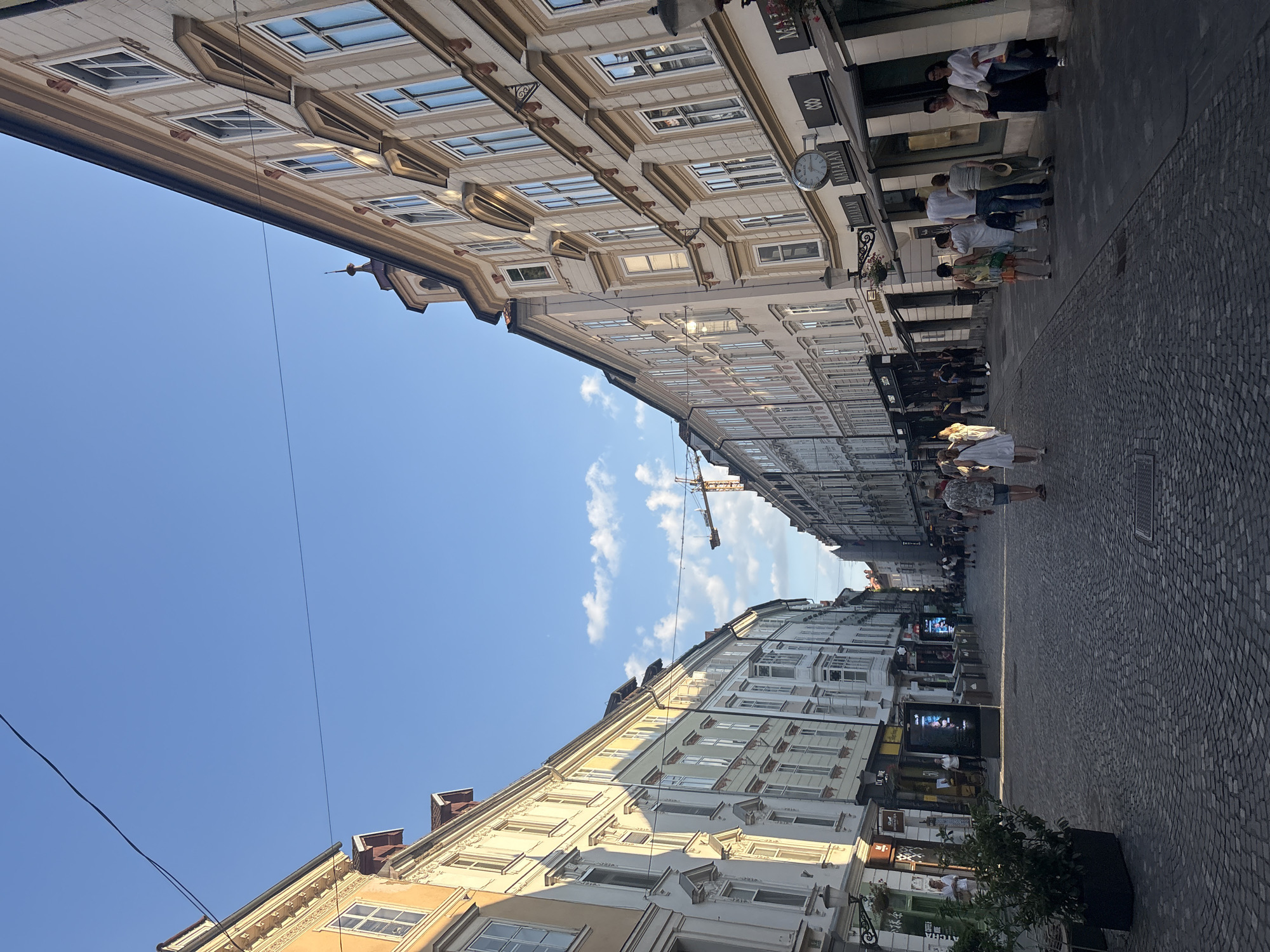 The image depicts a pedestrian street in a European city, likely lined with historical buildings. The architecture on both sides of the street is characterized by multi-story buildings with ornate facades, arched windows, and mansard roofs. The street is paved with cobblestones, and there are several people walking along the street. On the left side, there is a potted plant and some signage for businesses. On the right side, there are shops with awnings and display windows, including one with a visible clock. A construction crane is visible in the distance, suggesting ongoing development or renovation in the area. The sky is a clear blue with a few scattered clouds, and the lighting suggests it is late afternoon or early evening. There are overhead wires spanning across the street.