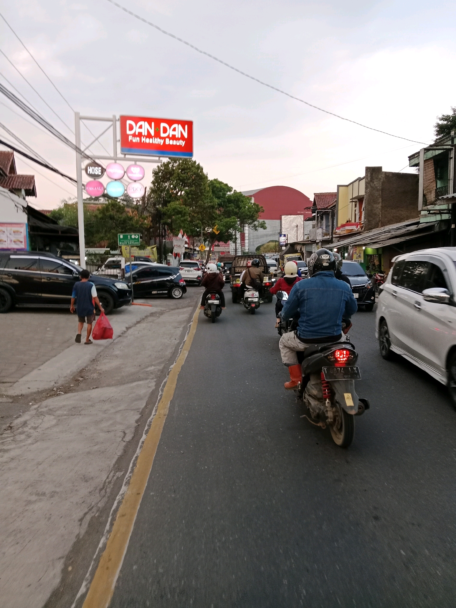 The image captures a street scene in Sirnagalih, Indonesia, characterized by moderate traffic and a mix of commercial and residential buildings. In the foreground, a person on a motorcycle, wearing a denim jacket and orange boots, is the primary subject. Their motorcycle's license plate is partially visible. To the left, a person in a blue shirt is walking on the sidewalk carrying a red bag. Several other motorcycles and cars are also present on the road, some moving and some parked.
A prominent sign for "DAN DAN Fun Healthy Beauty" is visible, accompanied by smaller illuminated circular signs below it, one labeled "HOSE." The architecture of the buildings suggests a bustling urban environment with varying building styles, including some with tiled roofs and visible electrical wiring overhead. The sky is overcast, indicating a cloudy or dusk setting. The road has a yellow line dividing the lanes. The overall impression is a typical day on a busy street in an Indonesian town.