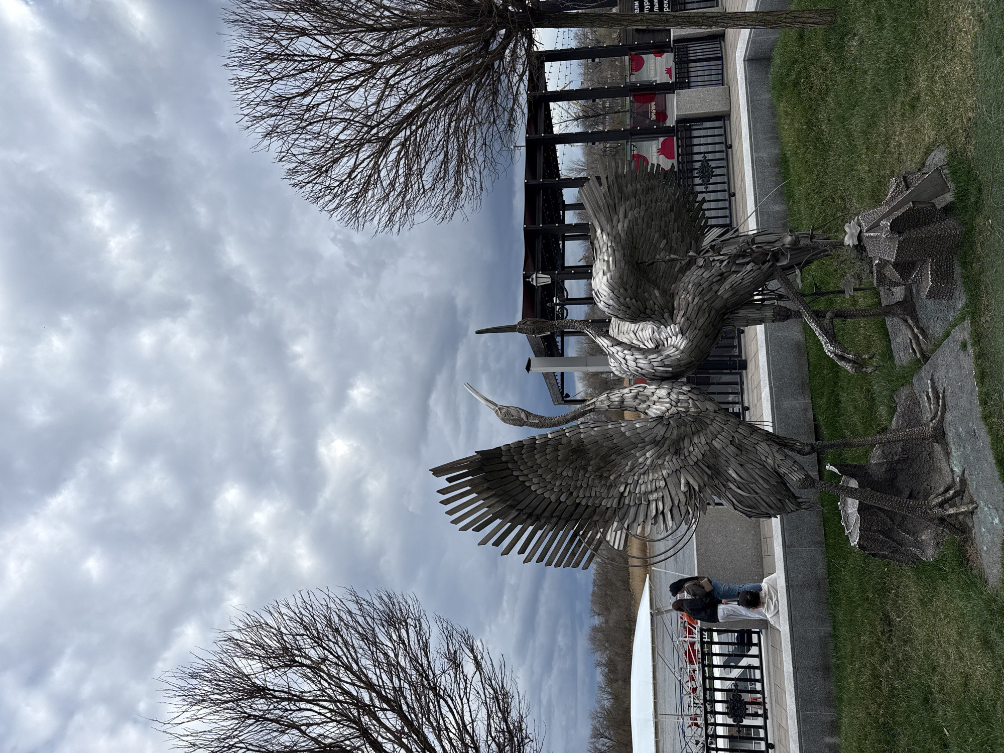 The image shows a sculpture of cranes in Rostov-on-Don, Russia. The sculptures are metal, depicting three cranes with outstretched wings. The sky is overcast. Bare trees are visible on either side of the frame. A fence runs behind the cranes. Two people are standing near the sculpture. A building with red accents is behind the fence. There is a patch of green grass to the right of the sculpture. The photograph is oriented vertically.