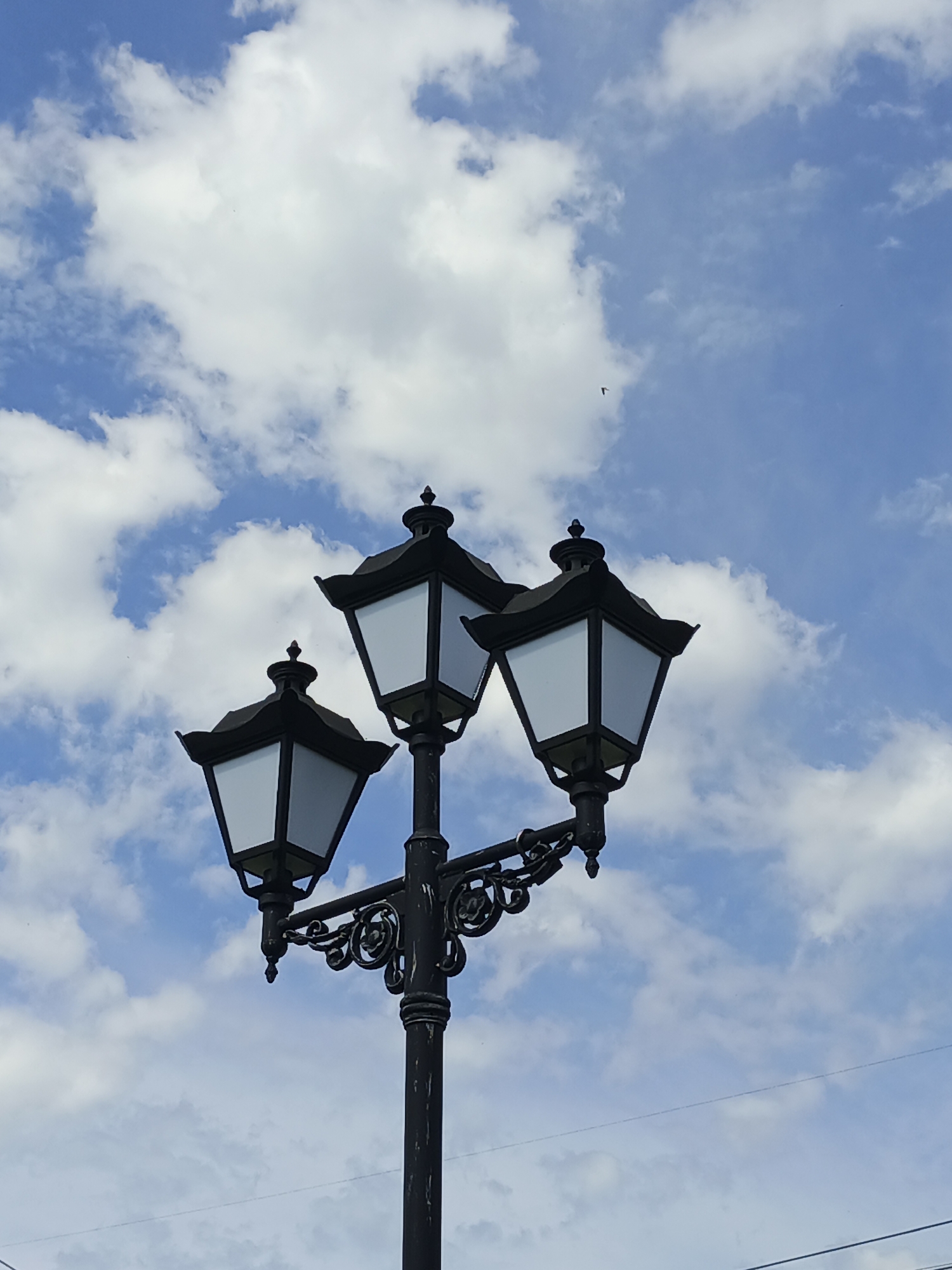 A black, three-headed lamppost stands against a bright blue sky dotted with fluffy white clouds. The lamppost has ornate decorative scrollwork on its arm. Each lamp housing is square with a pointed finial on top. The panels of the lamps are white and appear to be frosted or opaque. The sky takes up the majority of the frame. The scene is located in Zlatoust, Russia.
