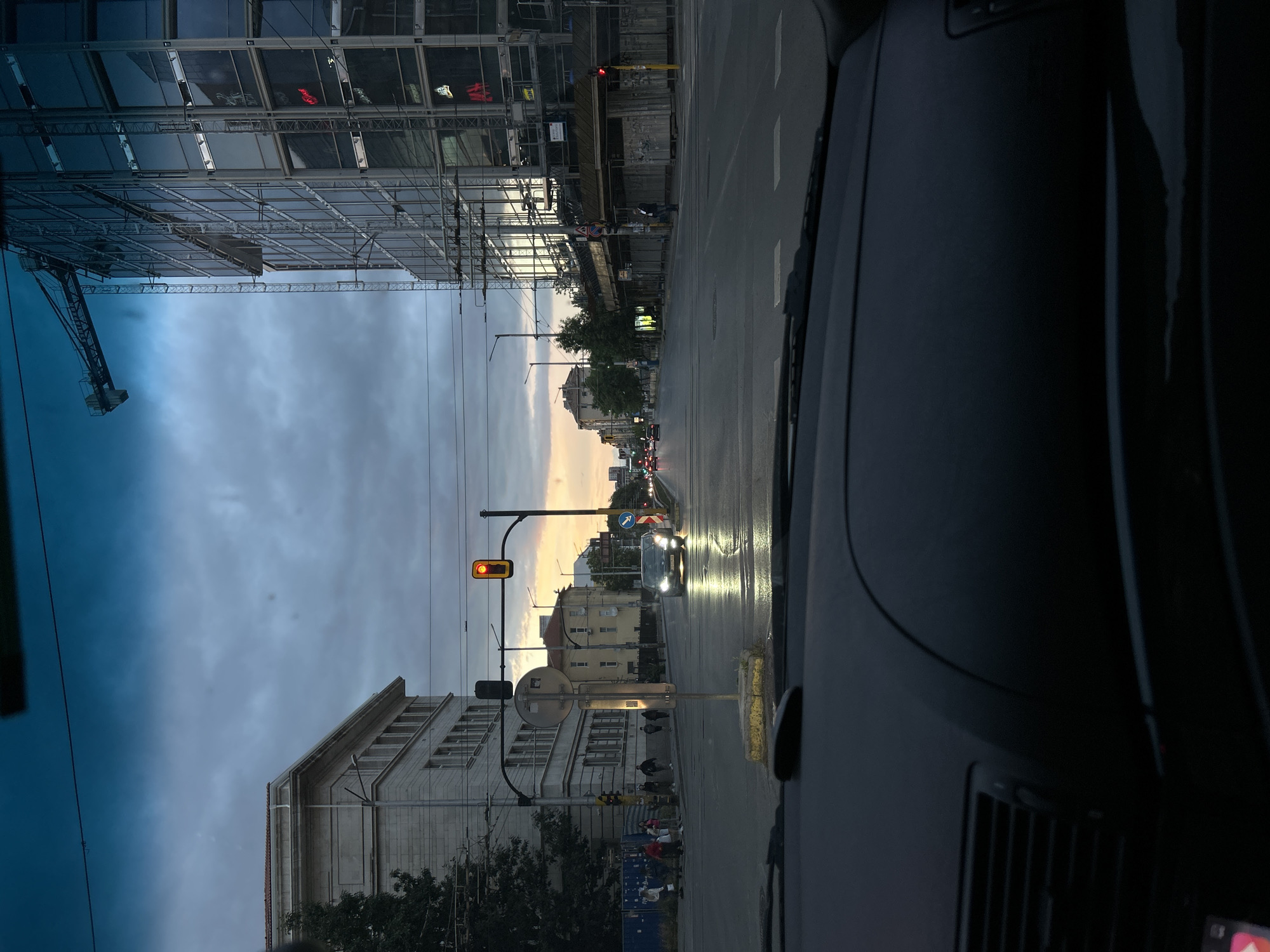 The image depicts a street scene at dusk, viewed from inside a car. The sky is overcast with a hint of sunset colors. On the left, a large, multi-story building with classical architectural elements stands beside a construction site with a crane. In the foreground, the car's dashboard is visible. The street is wet, indicating recent rain, and traffic lights are illuminated. Several vehicles and pedestrians are present on the street. The overall impression is that of an urban environment during the transition from day to night.