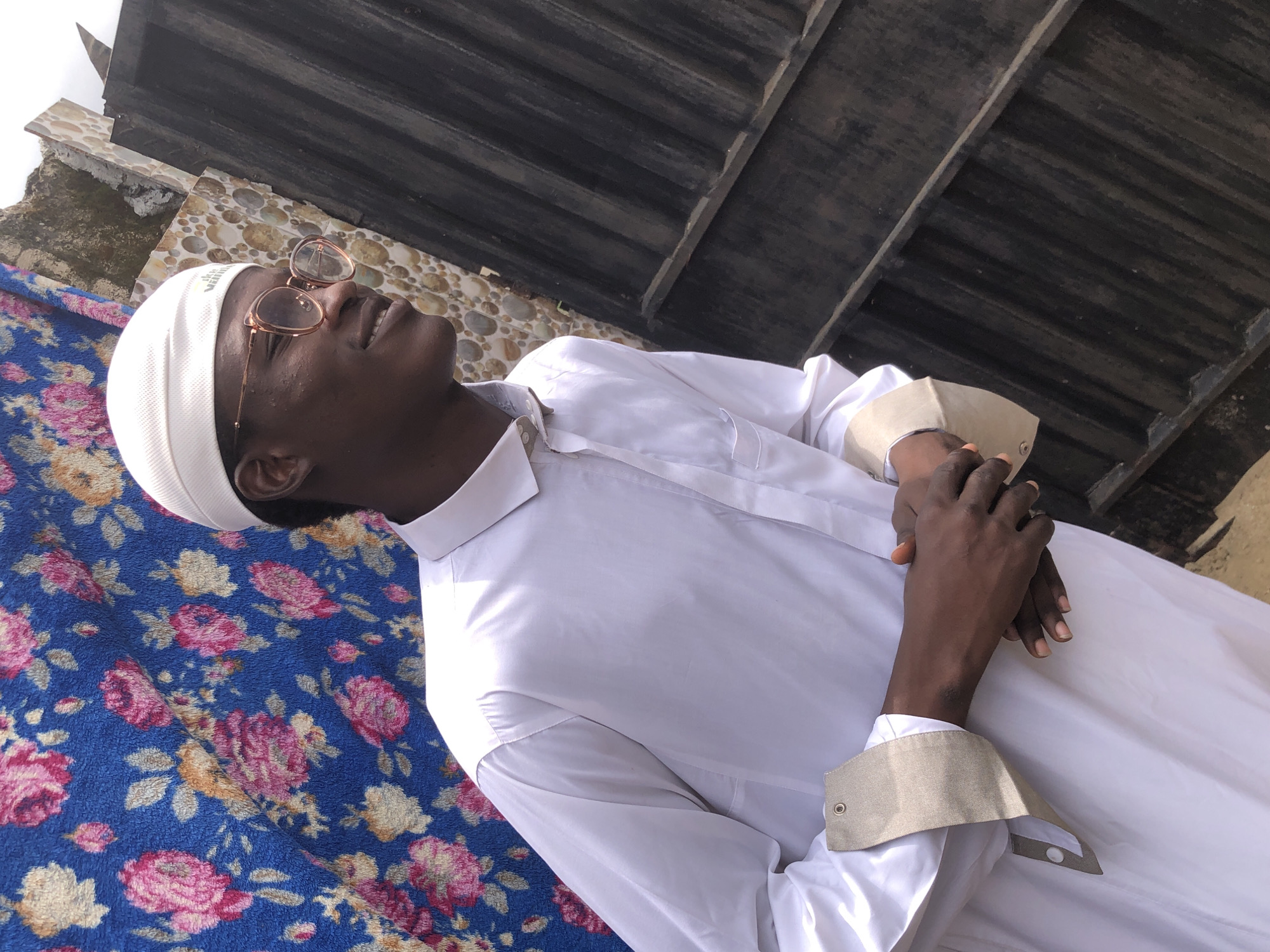 A medium shot captures a young Black man wearing a white kufi and a white thawb, standing in front of a floral blue fabric backdrop and a corrugated iron fence. He is looking to his right, with a slight smile. The thawb has a mandarin collar and a tan cuff with eyelets. He is wearing clear-rimmed glasses. His hands are clasped in front of him. The background consists of the patterned fabric on the left, and a wall with circular patterns and a dark, weathered corrugated iron fence on the right. The scene appears to be outdoors, possibly in a residential area of Ado, Nigeria, given the typical architecture and materials observed. The lighting suggests it is daytime, with some ambient light illuminating the man.