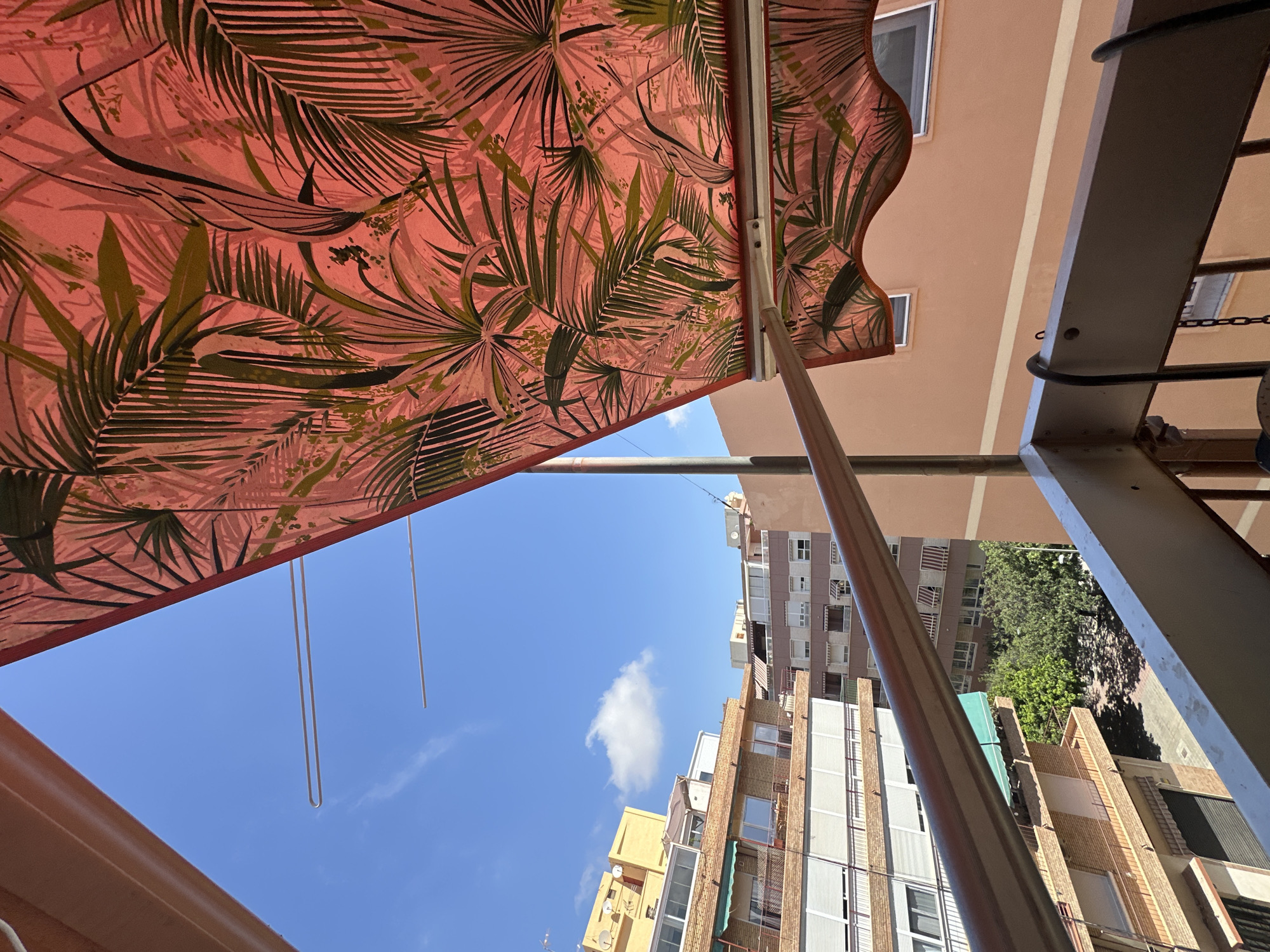 The image is taken from a balcony in an urban setting, looking up at a blue sky with a few clouds. A red awning with a tropical leaf pattern is partially extended, obscuring some of the sky and the surrounding buildings. The buildings are multi-story structures with visible balconies, windows, and awnings. In the distance, trees are visible in a courtyard or open space between buildings. The foreground shows a metal railing of the balcony. There are no people or animals visible in the image. The scene suggests a sunny day in a residential area.