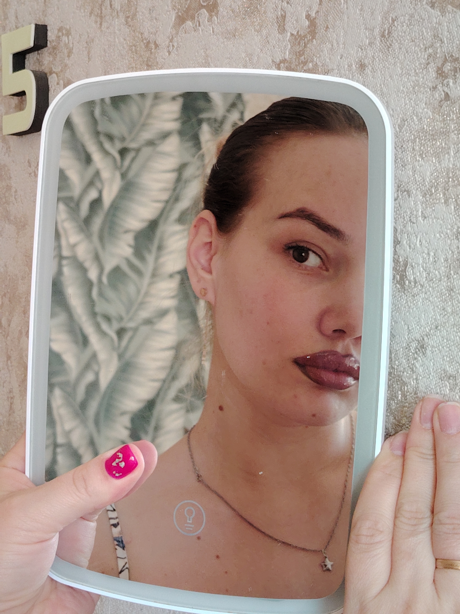 In an indoor setting within the city of Марковский, Russia, a person holds a white-framed, rectangular illuminated mirror with rounded corners. The mirror's reflective surface displays the right half of a female's face, characterized by dark hair pulled back, a brown eye, a defined eyebrow, a small stud earring in the right ear, and plump lips coated in dark, glossy lipstick. Visible on her neck is a thin chain necklace with a small star-shaped pendant, and a lightbulb icon, indicative of a touch control for illumination, is present on the mirror's lower edge. The background reflected in the mirror consists of wallpaper with a repeating pattern of light green tropical leaves. The individual holding the mirror is partially visible; the left thumb, adorned with bright pink nail polish featuring gold flake accents, is positioned on the left side of the mirror's frame. The right hand, with an unpolished nail and a plain gold-colored ring on the ring finger, grasps the mirror's right side. The physical background outside the mirror reveals a textured, beige-colored wallpaper, and a three-dimensional wooden or plastic number "5" with a dark border is affixed to the wall in the upper left corner of the frame.
