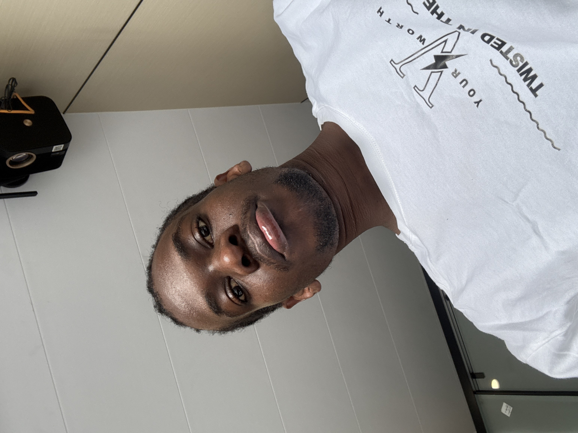 The image shows a low-angle, close-up selfie of a Black man wearing a white t-shirt. He has short, dark, curly hair and is looking directly at the camera with his lips slightly parted. The t-shirt has a graphic design with text that appears to read "YOUR WORTH IN THE TWISTED". In the background, there is a white paneled ceiling and a black electronic device mounted on the ceiling, possibly a projector or camera. The lighting suggests an indoor setting. There is no indication of the location being Yuzhong District, China.
