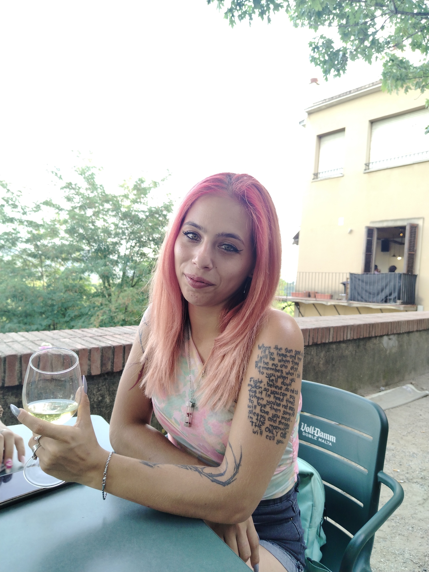 A woman with bright pink hair sits at a table outdoors, holding a glass of white wine. She is wearing a light-colored, tie-dye t-shirt and denim shorts. Her left arm is covered in tattoos, including a poem on her upper arm. She is sitting on a dark green plastic chair with the "Voll-Damm Doble Malta" logo visible. In the background, there is a low brick wall, lush green trees, and a light-colored building with windows. The scene suggests an outdoor dining or seating area, potentially in a city like Hostalric, Spain, given the architectural style and vegetation. The lighting indicates it is daytime, possibly late afternoon or early evening.