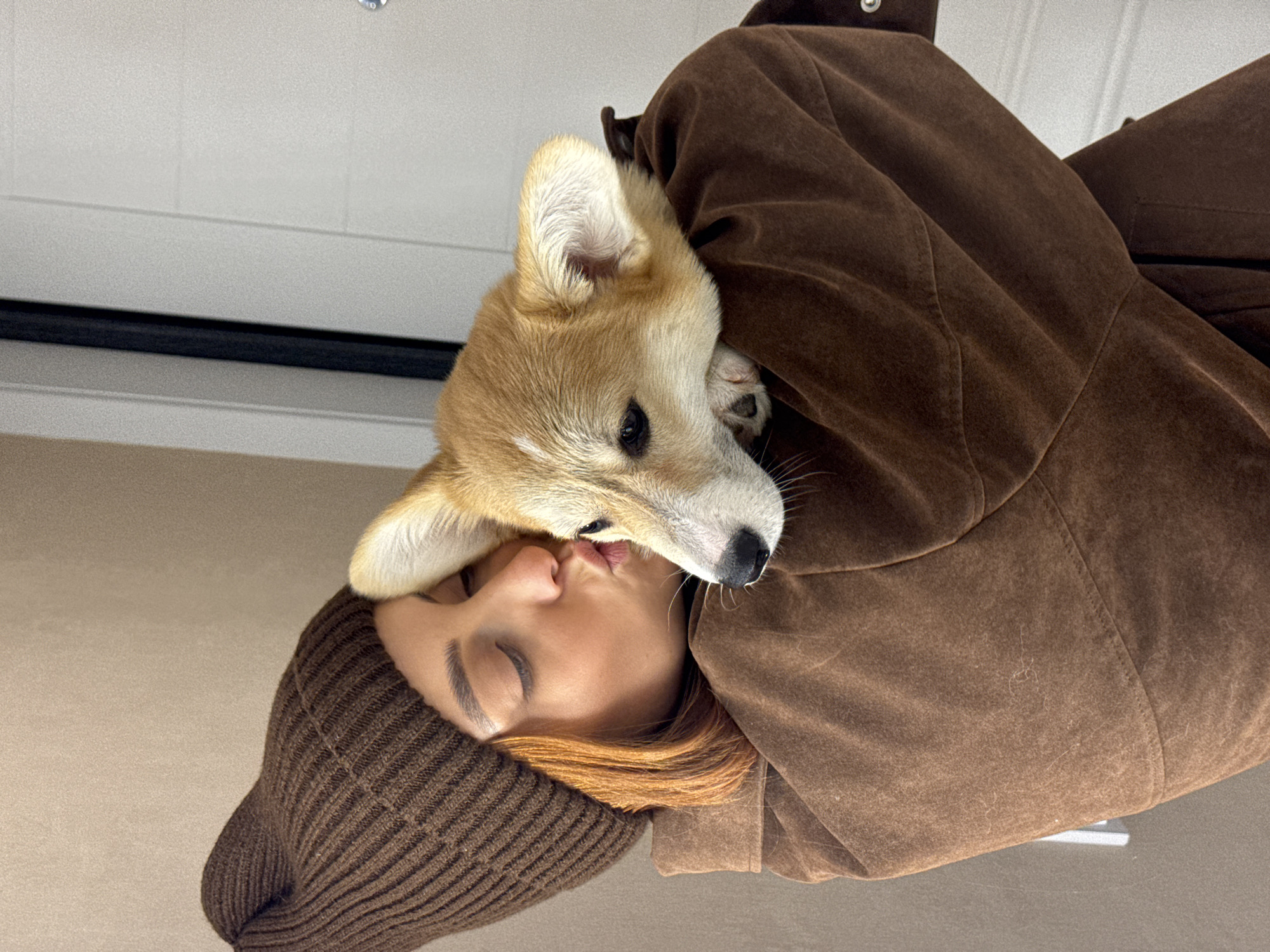 An indoor photograph depicts an adult female holding a Pembroke Welsh Corgi dog. The female, positioned with her left profile visible, wears a dark brown knit beanie and a textured dark brown jacket. Her reddish-brown hair is visible beneath the beanie. Her eyes are closed, and her lips are pursed towards the dog's head, indicating an affectionate gesture. The Corgi, a fawn and white dog with pointed ears and a black nose, rests its head on the female's left shoulder/arm. The background to the left consists of a plain light brown wall. To the right, a light-colored door with horizontal paneling and a dark vertical door jamb is visible, along with a metallic detail on the door. The scene takes place within a building in the city of Chaikovsky, Russia.