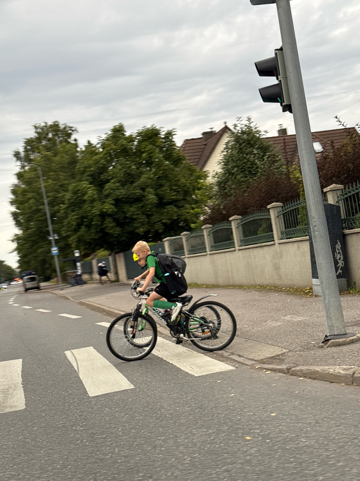 A young boy wearing a yellow helmet, a green t-shirt, black shorts, and green socks is cycling across a pedestrian crossing on an overcast day. He has a black backpack on. The bicycle is green and black. The pedestrian crossing is marked with white stripes on asphalt. To the right, a traffic light is visible, with the green light illuminated. Behind the crossing, there is a sidewalk with a low concrete wall and an ornate metal fence. Beyond the fence, houses with red-tiled roofs and trees can be seen. A street with a few cars is to the left of the crossing. The scene appears to be in an urban or suburban setting.