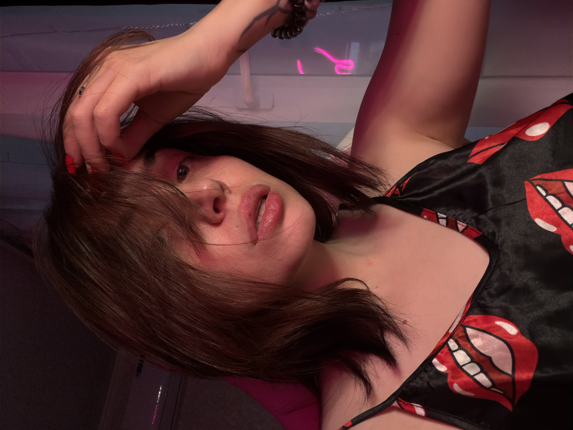 In an indoor, dimly lit setting, a young woman with dark brown hair is captured in a close-up shot, facing forward with her right hand touching her forehead. She is wearing a black camisole adorned with a repeating pattern of red lips with white teeth. The lighting creates a pinkish hue across her skin and surroundings. Her lips are slightly parted, and her eyes are partially obscured by her hair. The background is blurred, with hints of vertical blinds and neon pink light fixtures. The overall scene suggests a private and intimate moment.