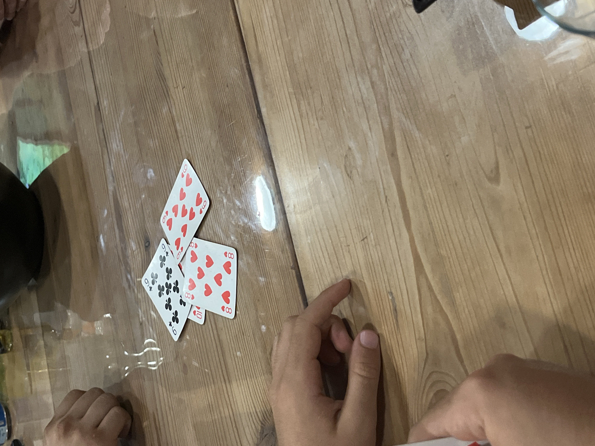 Three playing cards are spread on a wooden table. The cards are the 9 of Clubs, the 8 of Hearts, and the 10 of Hearts. A hand is visible on the left side of the frame, with fingers extended toward the table. The table surface is light brown with visible wood grain and some minor white debris. A partially visible black bowl and a glass are also on the table in the background. The scene appears to be indoors, with reflections on the table surface suggesting artificial lighting. No specific location in Kazanlak, Bulgaria can be identified from the image.