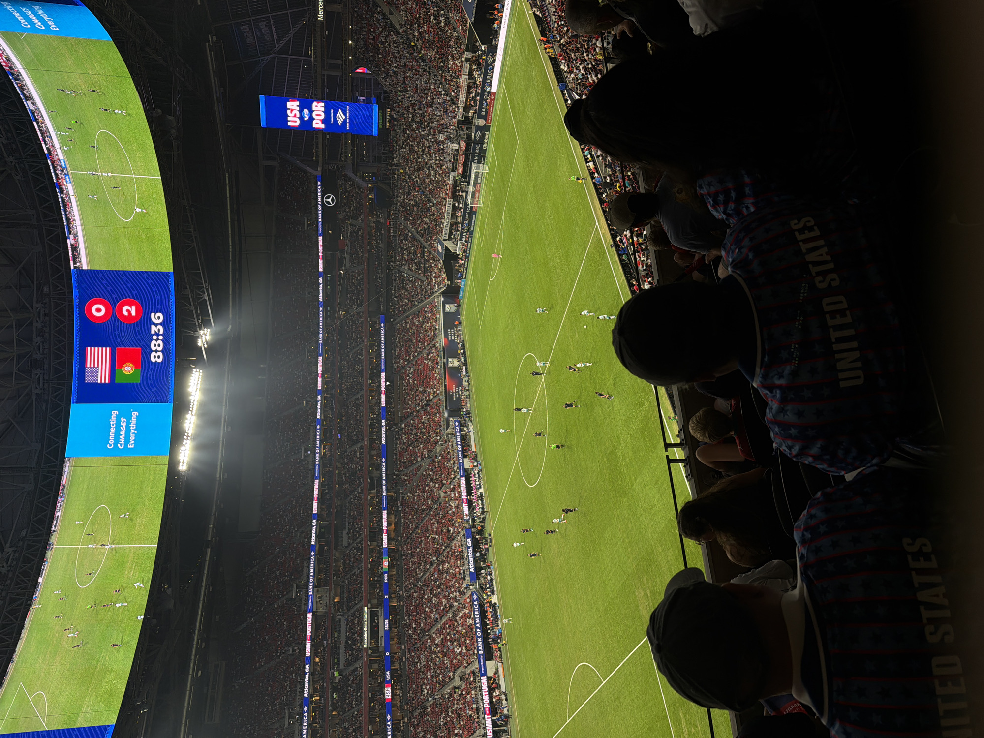 The image shows a soccer game in the Mercedes-Benz Stadium, located in Atlanta, United States of America. The game appears to be between the USA and Portugal, as indicated by the scoreboard. The score is 0-2 in favor of Portugal, and the game clock shows 88:36. A large crowd fills the stadium. In the foreground, portions of people wearing clothing with "United States" and stars on it can be seen. The stadium’s electronic signage, advertisements, and branding are visible.