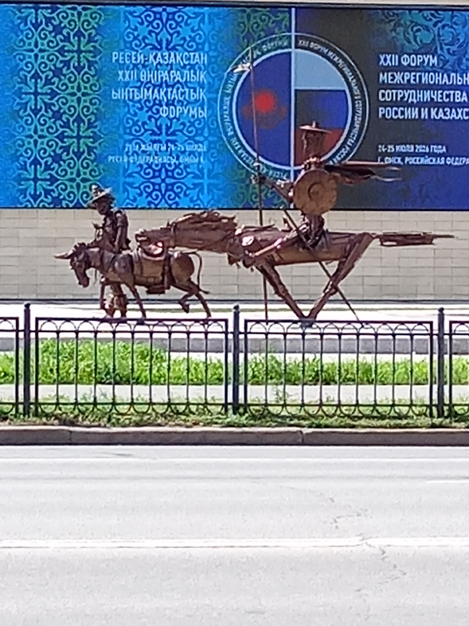 This image captures a bronze sculpture of a historical caravan, featuring a man riding a donkey loaded with goods, followed by another man on what appears to be a horse or a similar animal carrying more cargo. The sculpture is positioned in front of a large blue banner displaying text in Cyrillic, which reads: "XXII FORUM OF INTERREGIONAL COOPERATION BETWEEN RUSSIA AND KAZAKHSTAN" and "24-25 JULY 2020, OMSK, RUSSIAN FEDERATION". The banner also features a circular emblem divided into four quadrants, with the Russian flag in two of them and a red circle on a blue background in another. To the left of the banner, there is a decorative pattern of interwoven blue and green elements. A black metal fence with decorative scrollwork separates the sculpture and banner from a paved road in the foreground, with green grass visible behind the fence. The scene is set outdoors, under daylight conditions.