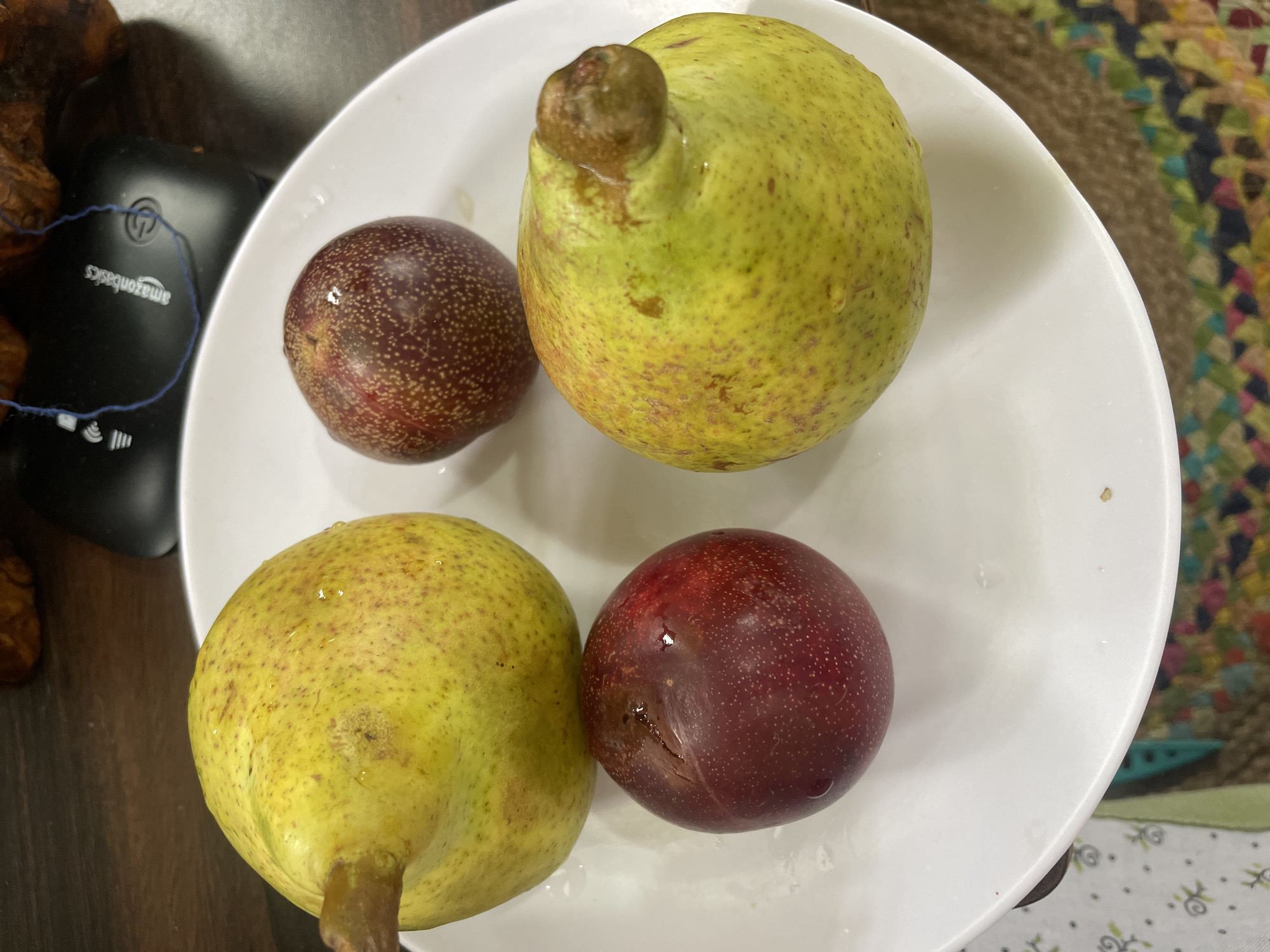 A white plate holds four pieces of fruit: two pears and two plums. The pears are a yellow-green color with a textured skin and a prominent stem at the top. The plums are a deep red-purple color, appearing smaller and rounder than the pears. The plate rests on a patterned surface, possibly a placemat or tablecloth, with a wooden surface visible in the background. A computer mouse and a tangled blue string are also present in the background, on the wooden surface. The lighting is natural, casting subtle shadows. The image does not provide any information to suggest its location in Bhagsu, India.