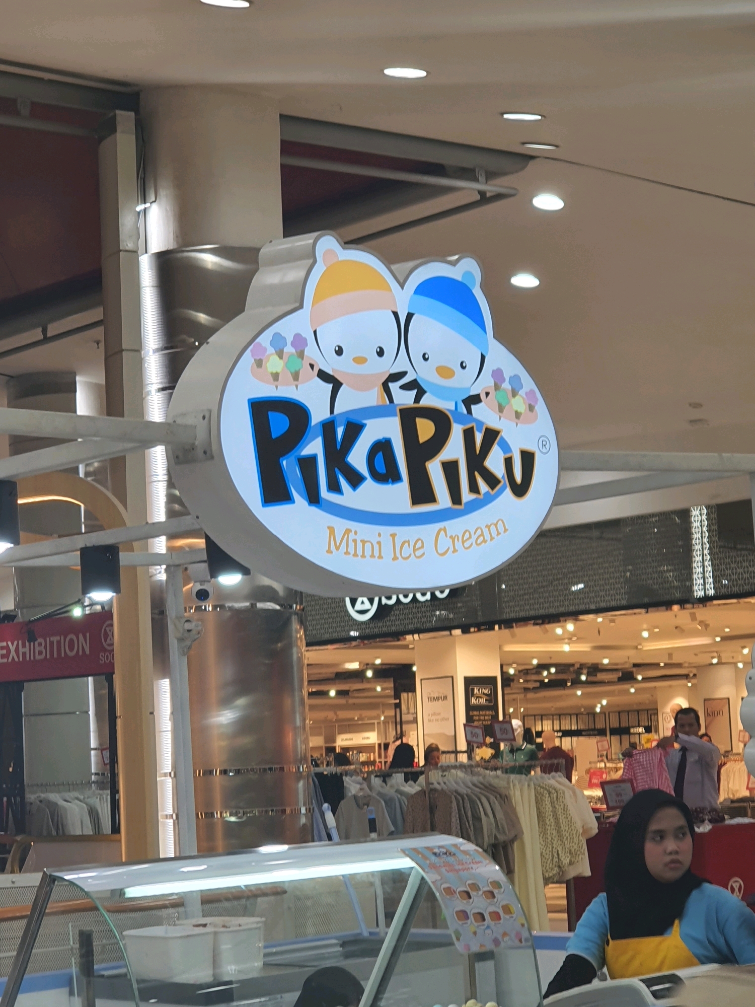 The image depicts an interior scene, likely within a shopping mall, with a prominent sign for "Pika Piku Mini Ice Cream" hanging overhead. The sign features cartoon penguins wearing beanies and holding ice cream cones. Below the sign, an ice cream display case is visible, with a person wearing a hijab and a blue and yellow uniform standing behind it. In the background, various clothing racks and store displays are visible, along with other shoppers and mall staff. The lighting suggests an indoor environment with artificial illumination from overhead lights and the store displays. The context of "North Jakarta, Indonesia" places this scene within a specific geographic and cultural setting.