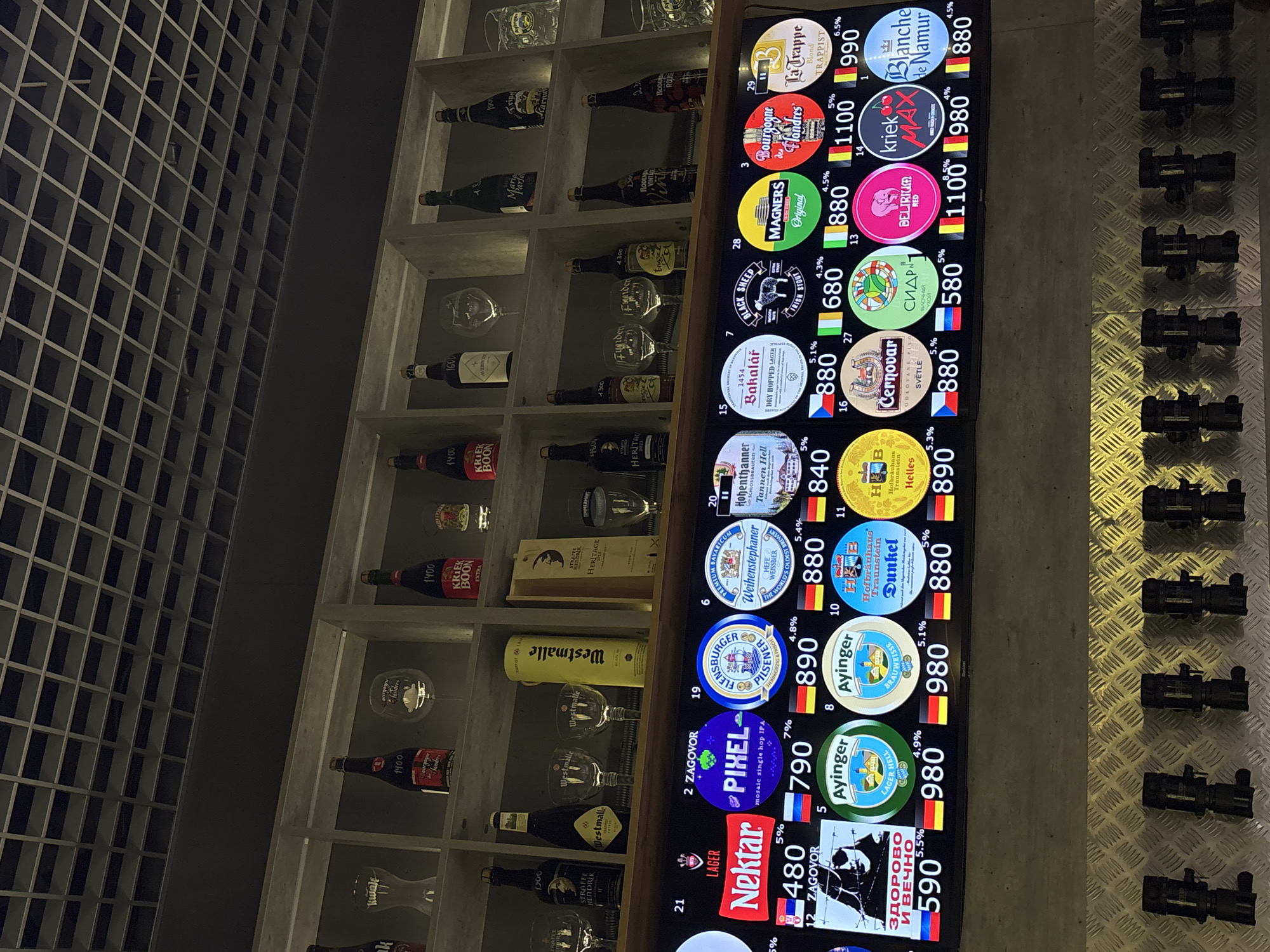 The image shows a bar setting with a wall of shelved beer bottles and glasses on the left and a column of beer taps with a digital display on the right. The display shows various beer brands with their names, logos, alcohol percentages, and prices (presumably in Russian rubles). Some brands visible on the display include "Nektar," "Pixel," "Ayinger," "Flensburger," "Weihenstephaner," "Hofbräuhaus Traunstein," "Hohenthanner," "Bakalář," "Cernovar," "Magners," "Bourgogne des Flandres," "La Trappe," "Delirium Red," "Kriek Max," and "Blanche de Namur." The location is potentially a bar or restaurant offering a wide selection of beers, specifically in Vnukovo, Russia (implied from OCR of "ЗДОРОВО И ВЕЧНО"). No people are visible in the image.