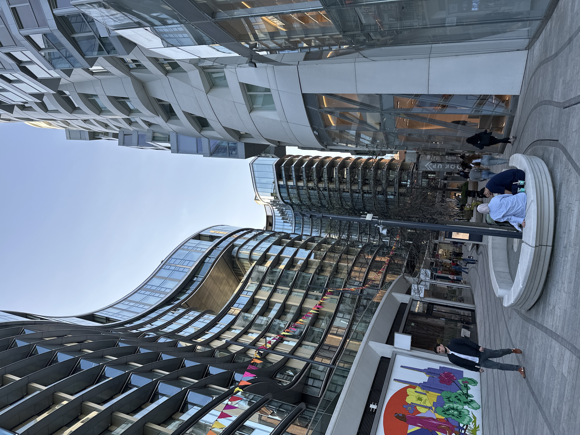 The image depicts an urban scene in London, United Kingdom, featuring modern architectural buildings. A man in a suit stands in the foreground beside a large, colorful mural on a wall. Behind him, a group of people interacts around a semicircular structure, possibly a bench or fountain. The architecture is characterized by curved lines and glass facades, with decorative pennants hanging from one building. The sky is clear, suggesting daylight.