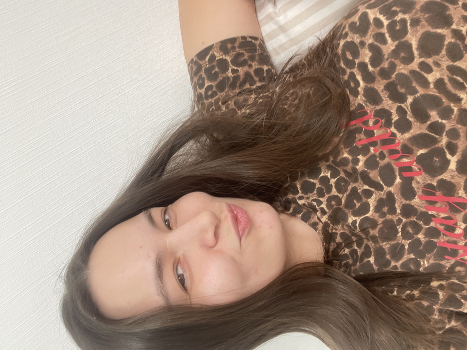 A woman with long brown hair poses lying on a bed. She is wearing a brown leopard print shirt with red writing on the front. The background includes a white wall and the edge of a striped pillow. Her face is turned toward the camera with a slight smile. The photo is rotated.