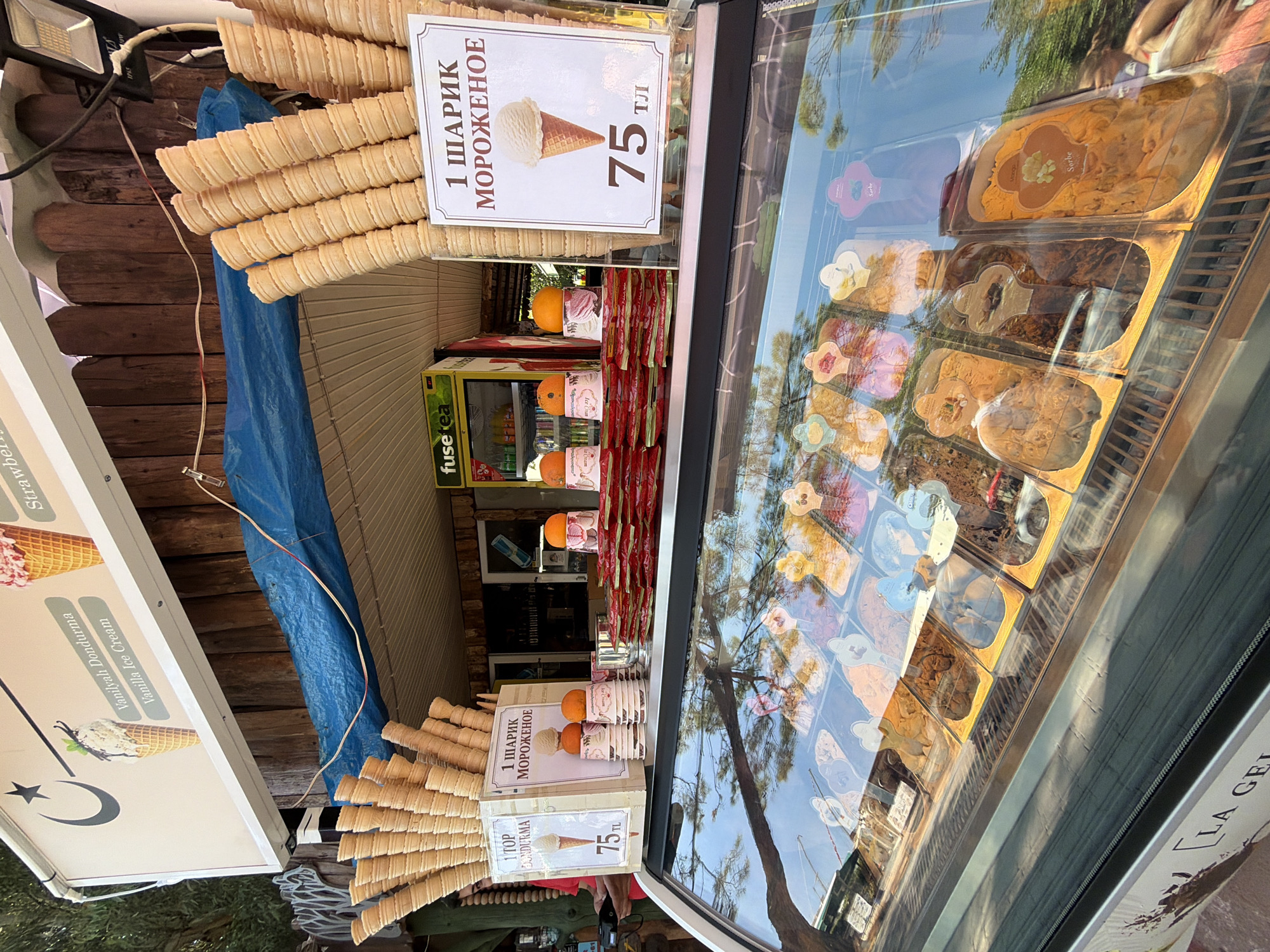 The image captures an outdoor ice cream stand in Kemer, Turkey, on a sunny day. In the foreground, a glass display case showcases various flavors of gelato and sorbet, with labels like "Mango Sorbet" visible. Above the display, a sign advertises "1 ШАРИК МОРОЖЕНОЕ" (1 scoop of ice cream) for 75 TL, featuring an image of an ice cream cone. Another sign on the left advertises "1 TOP DONDURMA" (1 top ice cream) for 75 TL, with an image of vanilla ice cream. Stacks of empty ice cream cones are prominently displayed. Behind the counter, a small refrigerator labeled "fuse tea" is visible, along with oranges placed on top of some ice cream cups. The background consists of wooden structures and some greenery, suggesting an outdoor setting. No people are clearly visible in a discernible action within the frame, though a person's arm and hand holding a device are partially visible on the left side of the image.