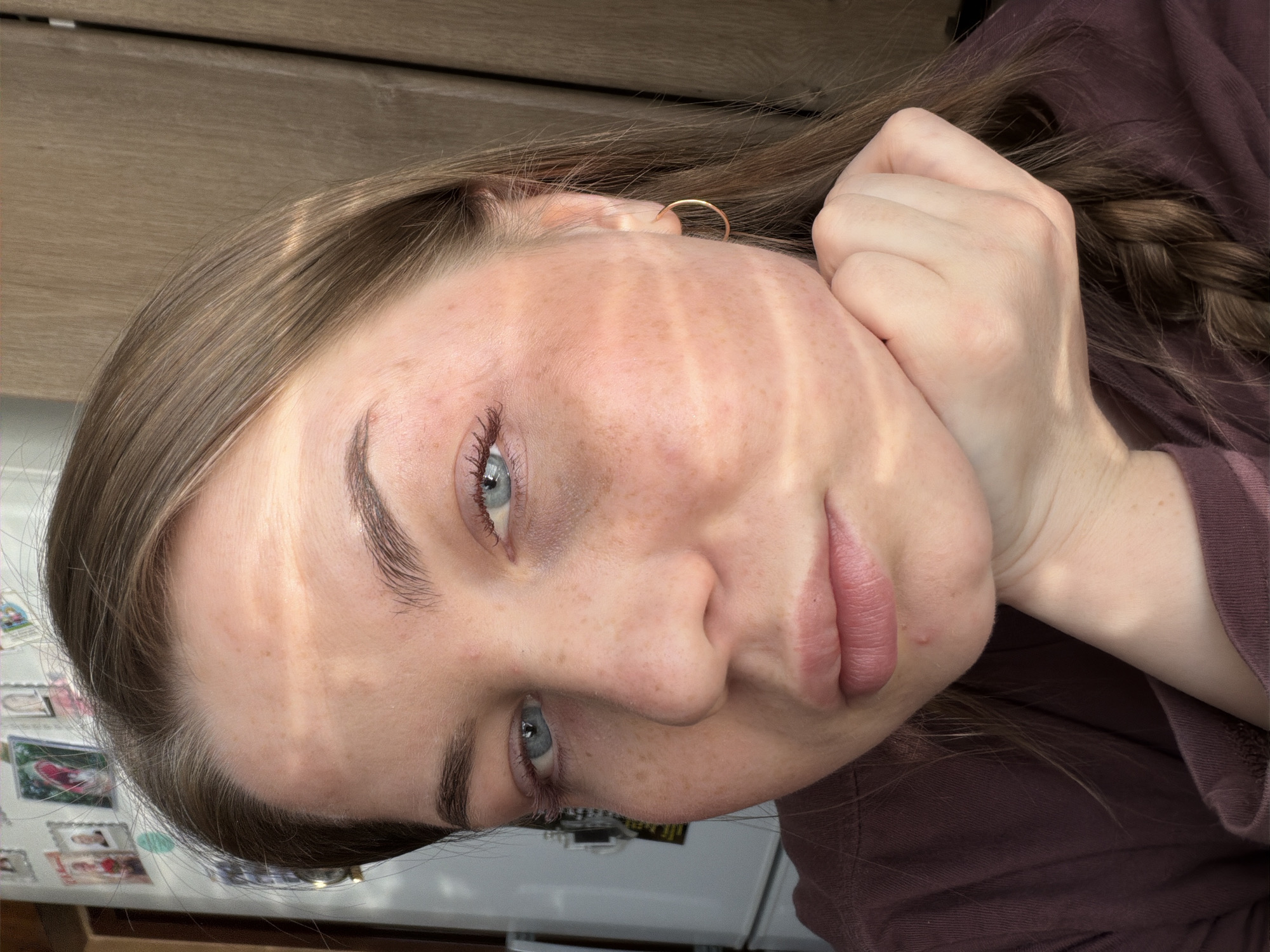A close-up, angled portrait shows a Caucasian woman with light brown hair and blue eyes. She is wearing a brown long-sleeved shirt. A gold hoop earring is visible in her left ear. Her right hand is held near her face, with her hair braided and resting against her hand. The face is freckled, and shadows from what appears to be blinds are cast across the face. The background includes a wooden cabinet and a white refrigerator covered with pictures or magnets. The location in the city of Уральское, Russia, cannot be determined from the image.