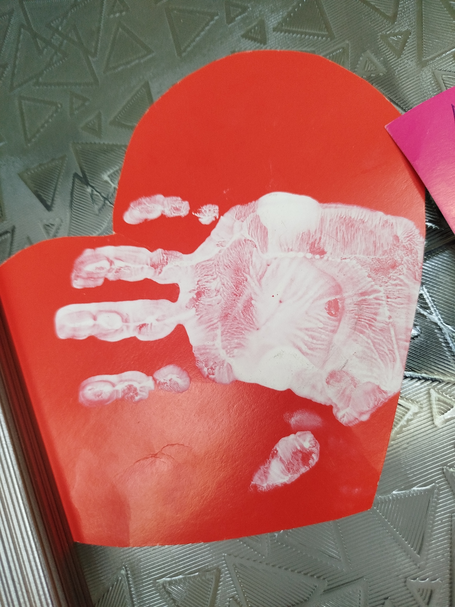 A close-up, top-down view captures a red heart-shaped piece of paper with a white handprint on it. The handprint, likely from a child, is partially smudged, with distinct imprints of fingers and the palm. The paper rests on a textured, grey surface with a repeating triangular pattern. To the right, a portion of a pink card is visible. The image appears to be taken indoors. The location of Крывасельскі сельскі Савет, Belarus is not discernible from the visual information provided in the image.