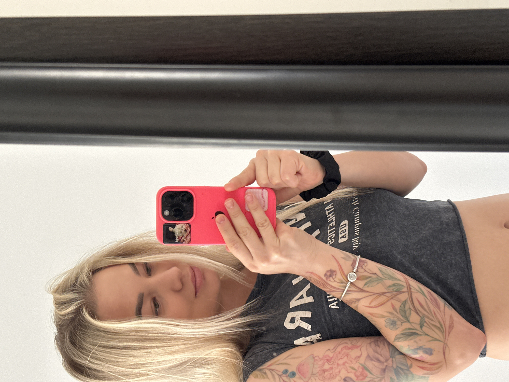 A blonde woman is captured in a mirror selfie taken indoors in Budva, Montenegro. She is holding a smartphone with a bright pink case in her hands, positioned to photograph her reflection. Her long, straight blonde hair cascades around her face as she looks down towards the phone.
She is wearing a grey cropped t-shirt, exposing her midriff, and a black scrunchie is on her right wrist. Her arms are extensively decorated with colorful, floral, and nature-themed tattoos, including a jellyfish design on her right forearm. A silver bracelet with a prominent clear gem adorns her left wrist.
The text on her t-shirt, visible as a reflection, appears as "ADDA... ZOTITJHTA...", which, when unmirrored, likely spells out "ATHLETIC...". On the back of her phone case, a small, mirrored sticker is visible, with text starting "AAAAAV..." that is difficult to fully decipher. The background of the reflection is simple, featuring a dark horizontal bar positioned above a plain light-colored wall, possibly a shelf or part of the mirror frame.